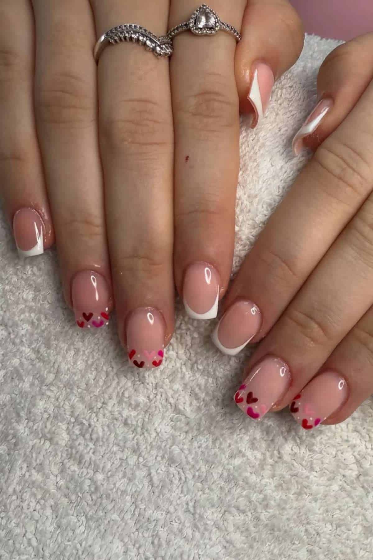 Two hands with manicured nails; one hand has classic French tips, the other has pink tips with small red and pink heart designs. Both hands rest on a white towel.