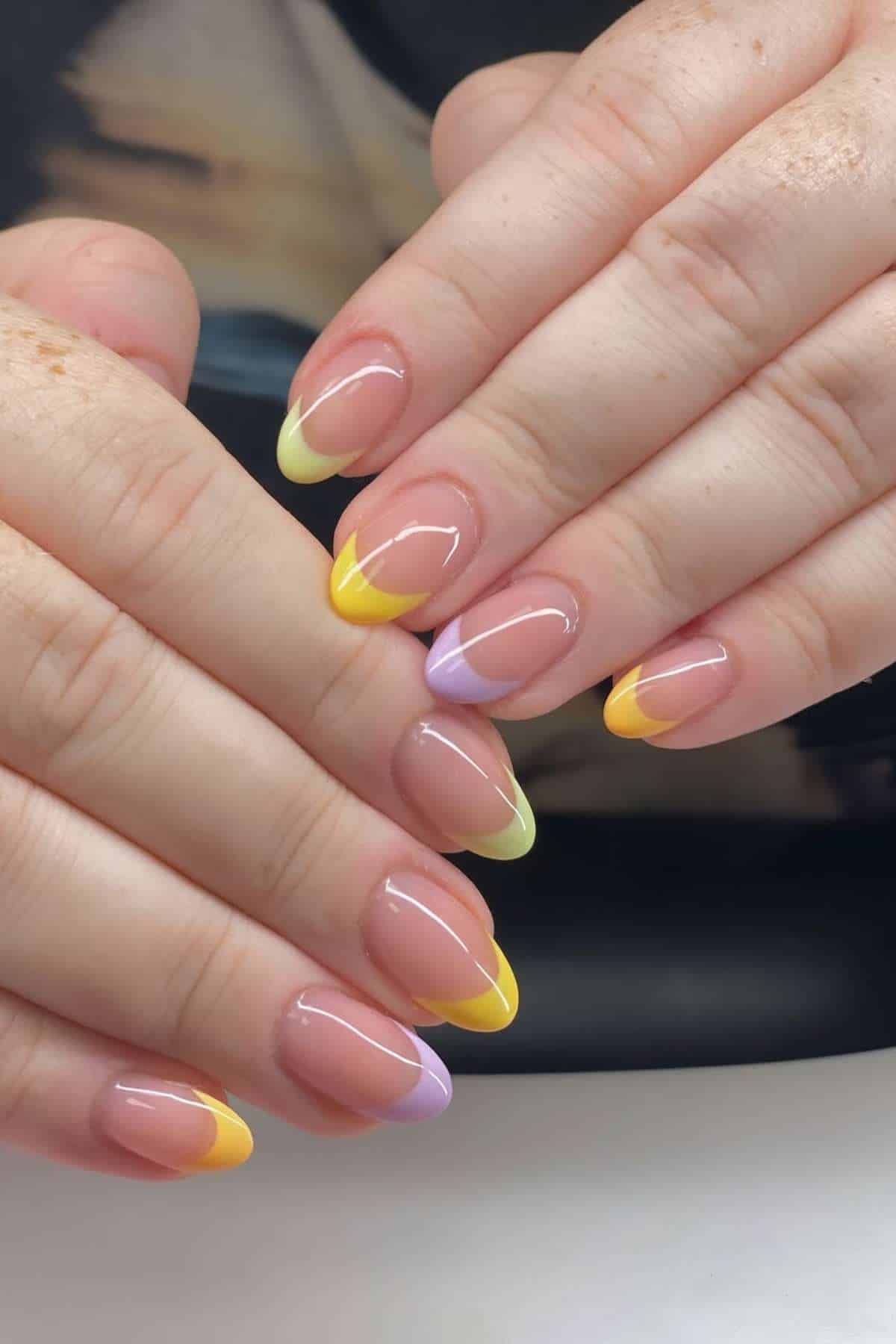 Close-up of hands with almond-shaped nails featuring a French tip design in yellow and lavender on a natural base.