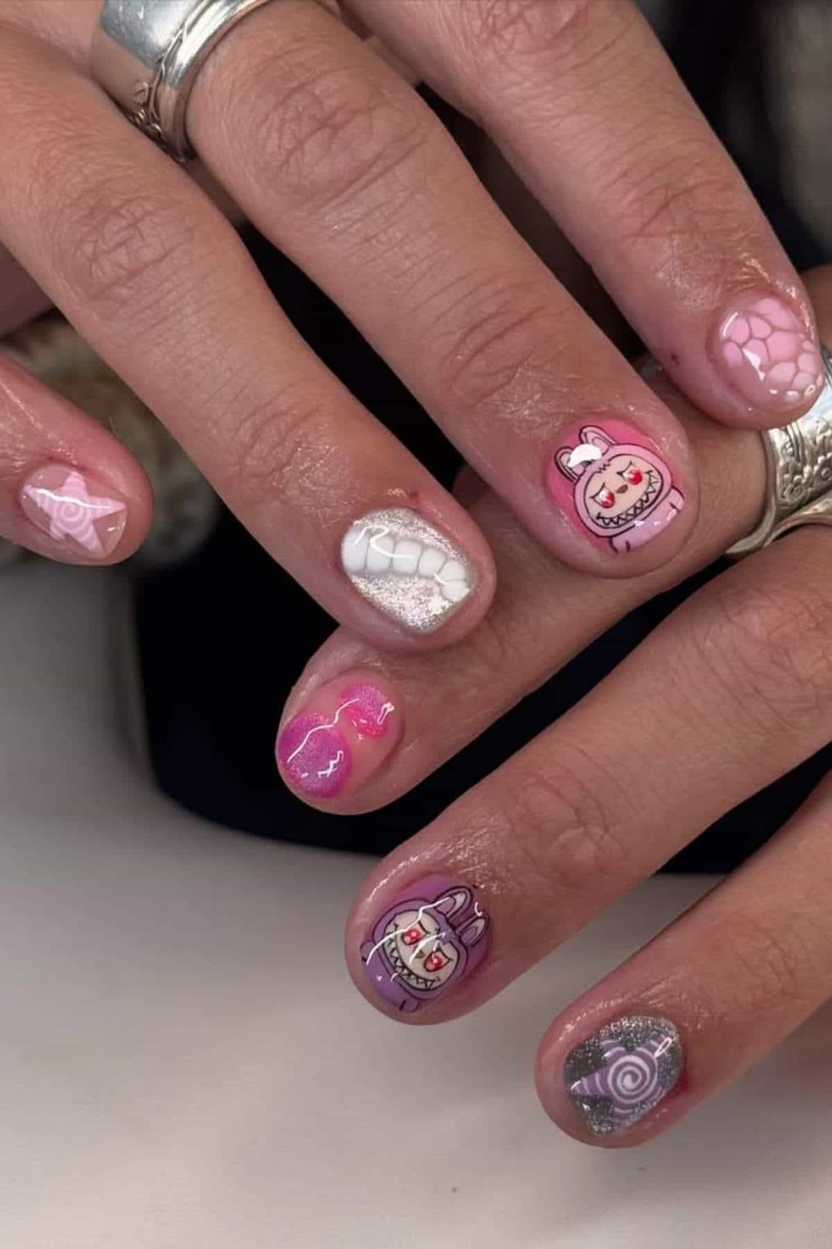 A hand with short nails painted in pink, purple, and white shades, featuring cartoon faces, swirls, and heart designs. The person is wearing silver rings.