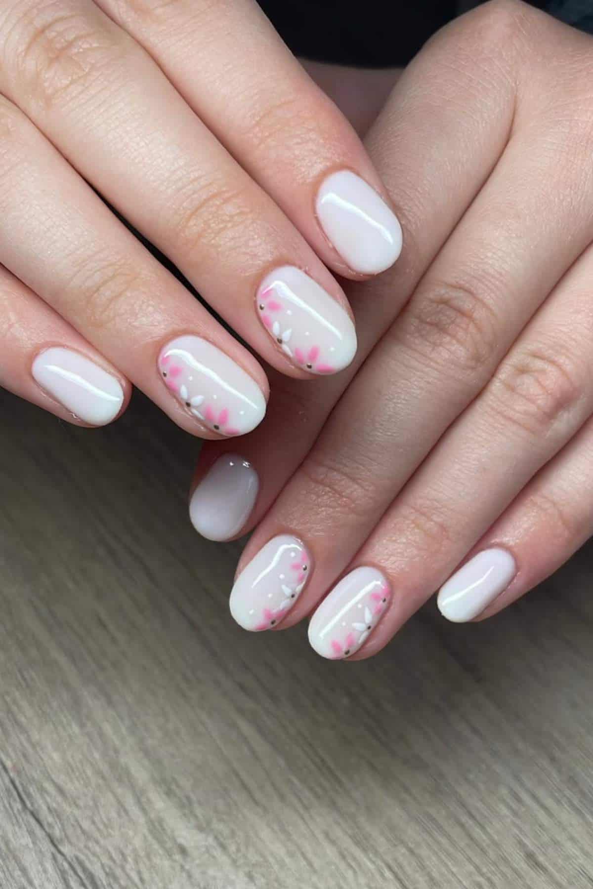 A close-up of hands with short, oval-shaped nails painted pale pink and decorated with small pink and green floral designs on some nails.