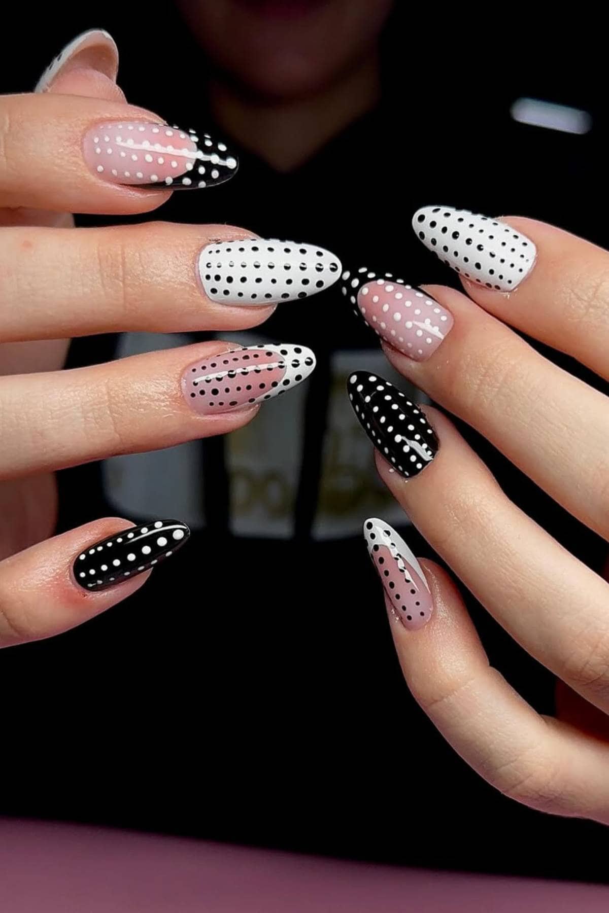 Hands with almond-shaped nails painted in black, white, and pink, each nail featuring a pattern of white or black polka dots.
