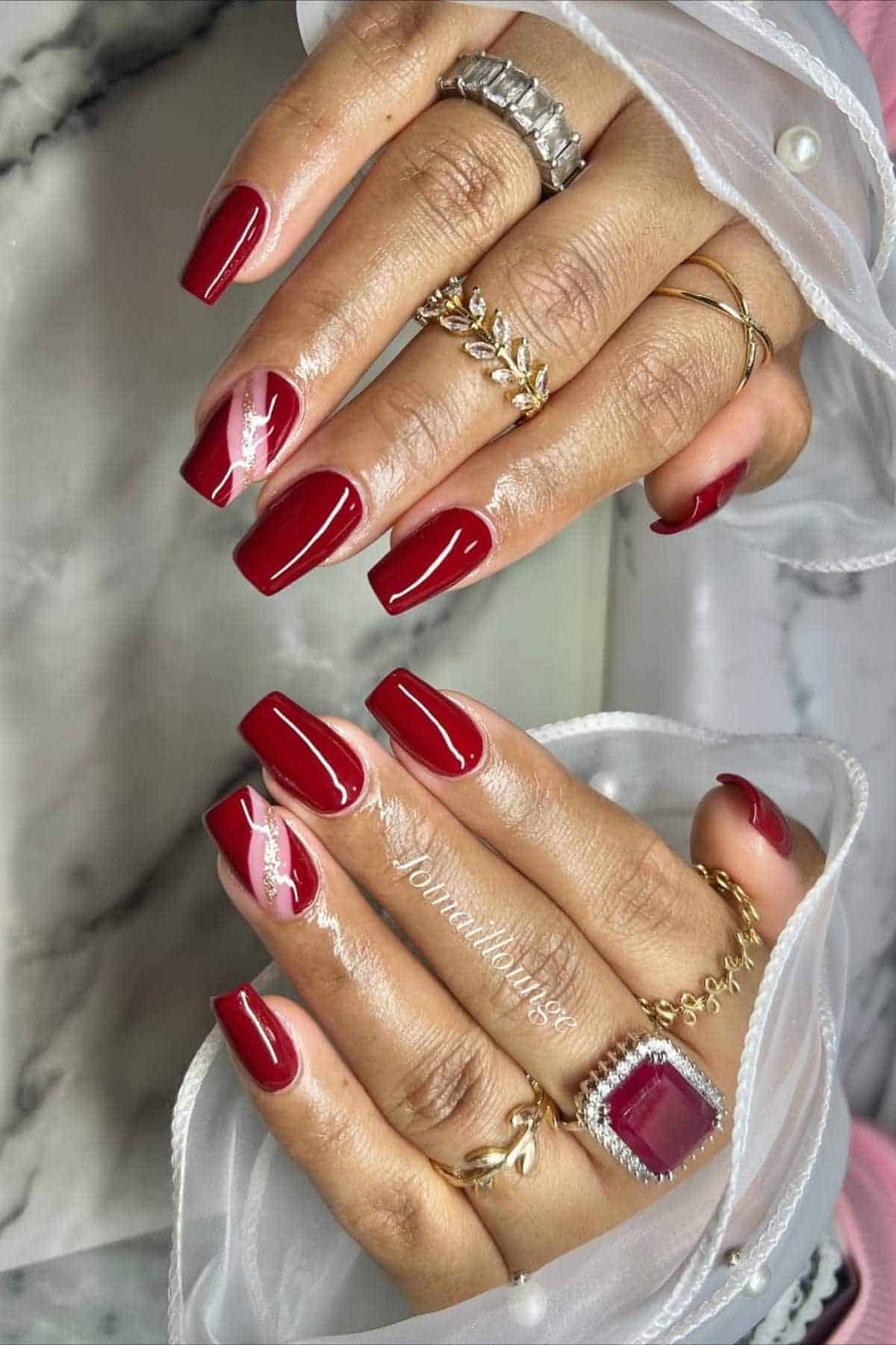 Hands with long, square-shaped red manicured nails, accented with light swirl designs, wearing multiple gold and silver rings on marble background.