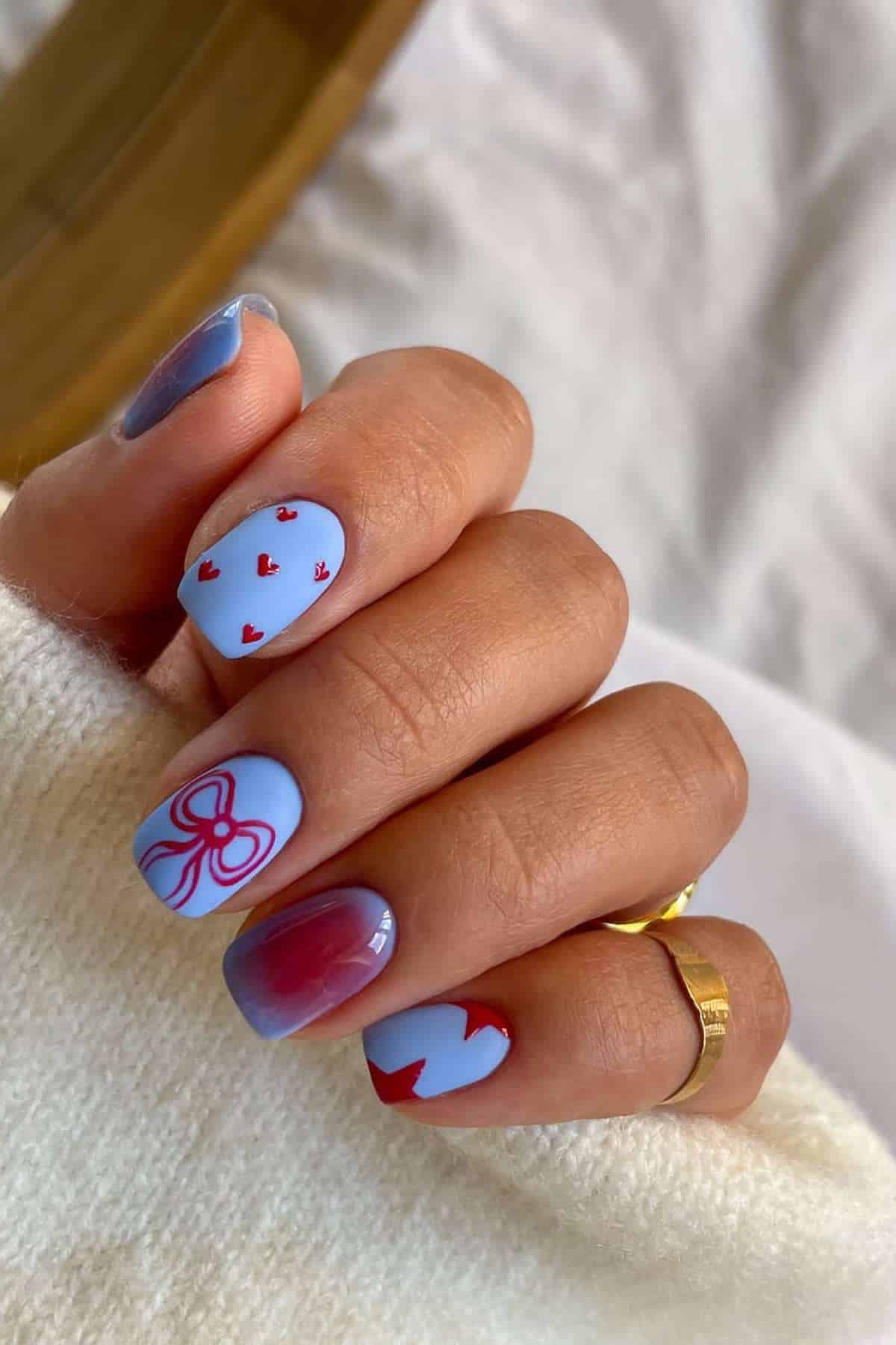 A hand with short nails painted in pastel purple and blue, decorated with small red hearts, a red bow, a zigzag pattern, and a red-to-purple gradient design.