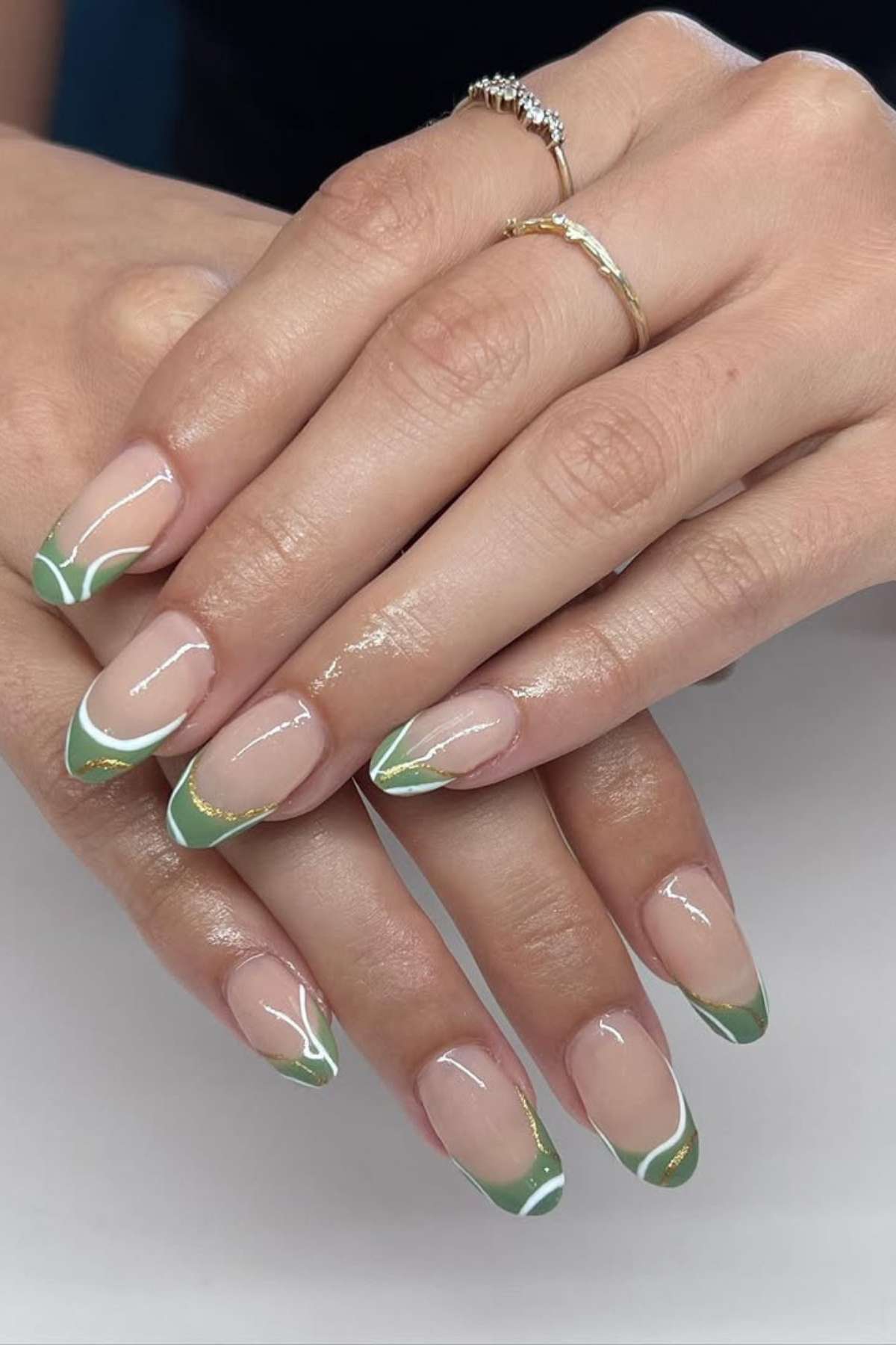Hands with manicured nails featuring green and white French tips and gold accent lines; wearer has two gold rings on ring and middle fingers.
