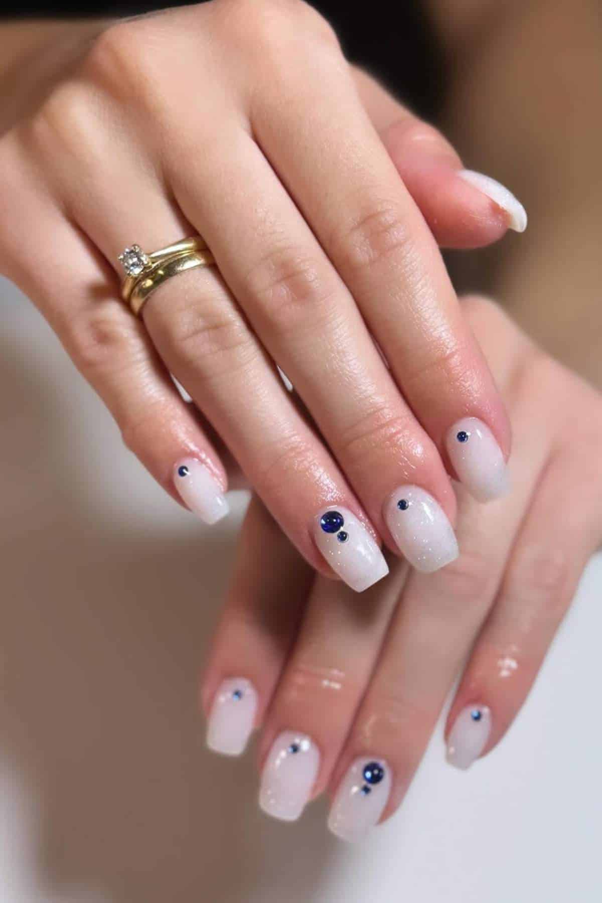 Hands with light pink, glossy, square-shaped nails decorated with small blue gems. The person is wearing two gold rings with a clear stone on the ring finger.