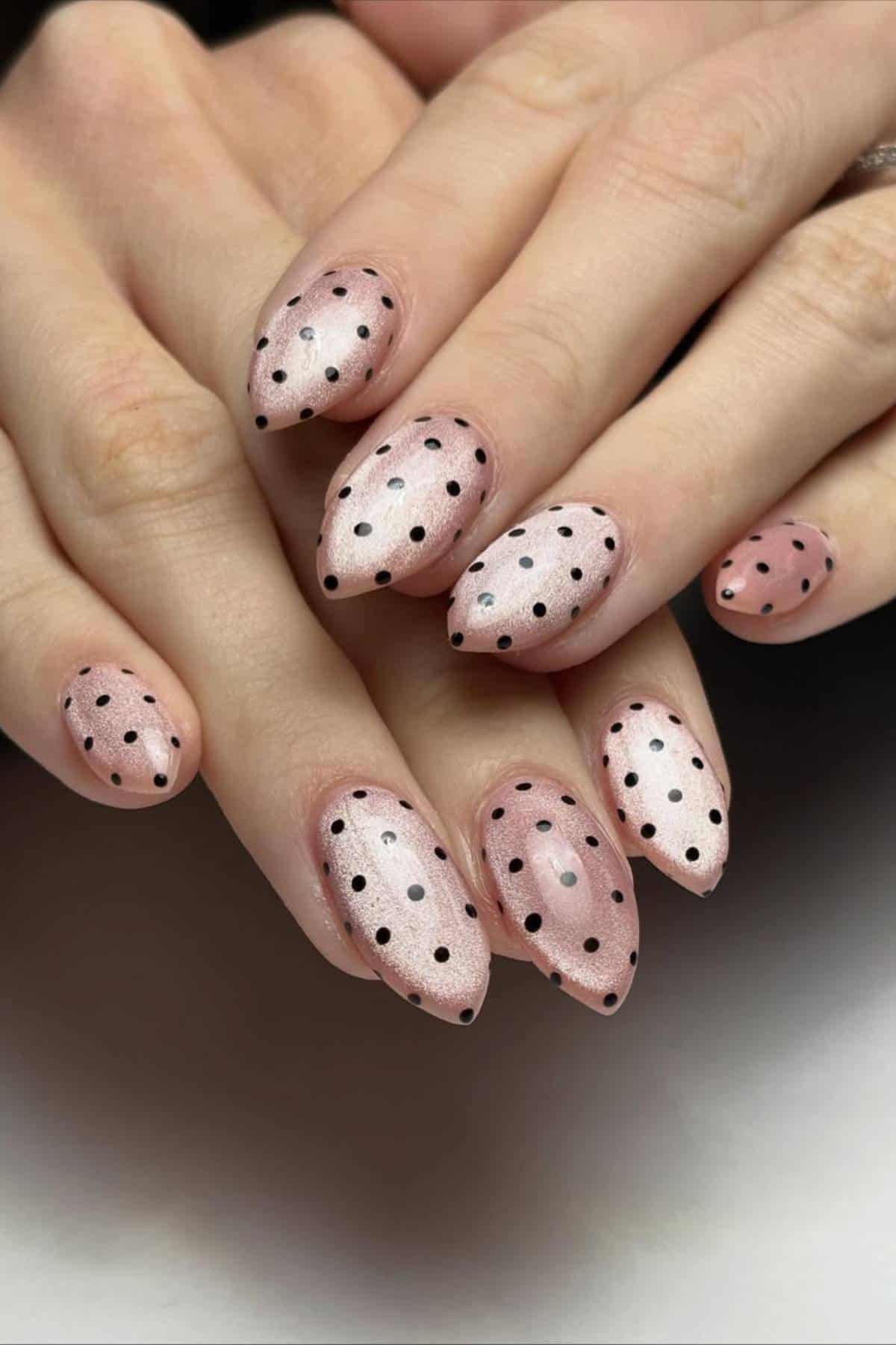 Hands with almond-shaped nails painted in a shimmery pink polish, each nail decorated with evenly spaced black polka dots.