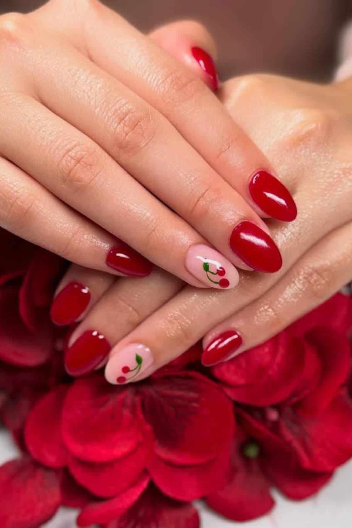 Hands with almond-shaped red nails; one nail on each hand is light pink with a small cherry design. Red flowers are in the background.