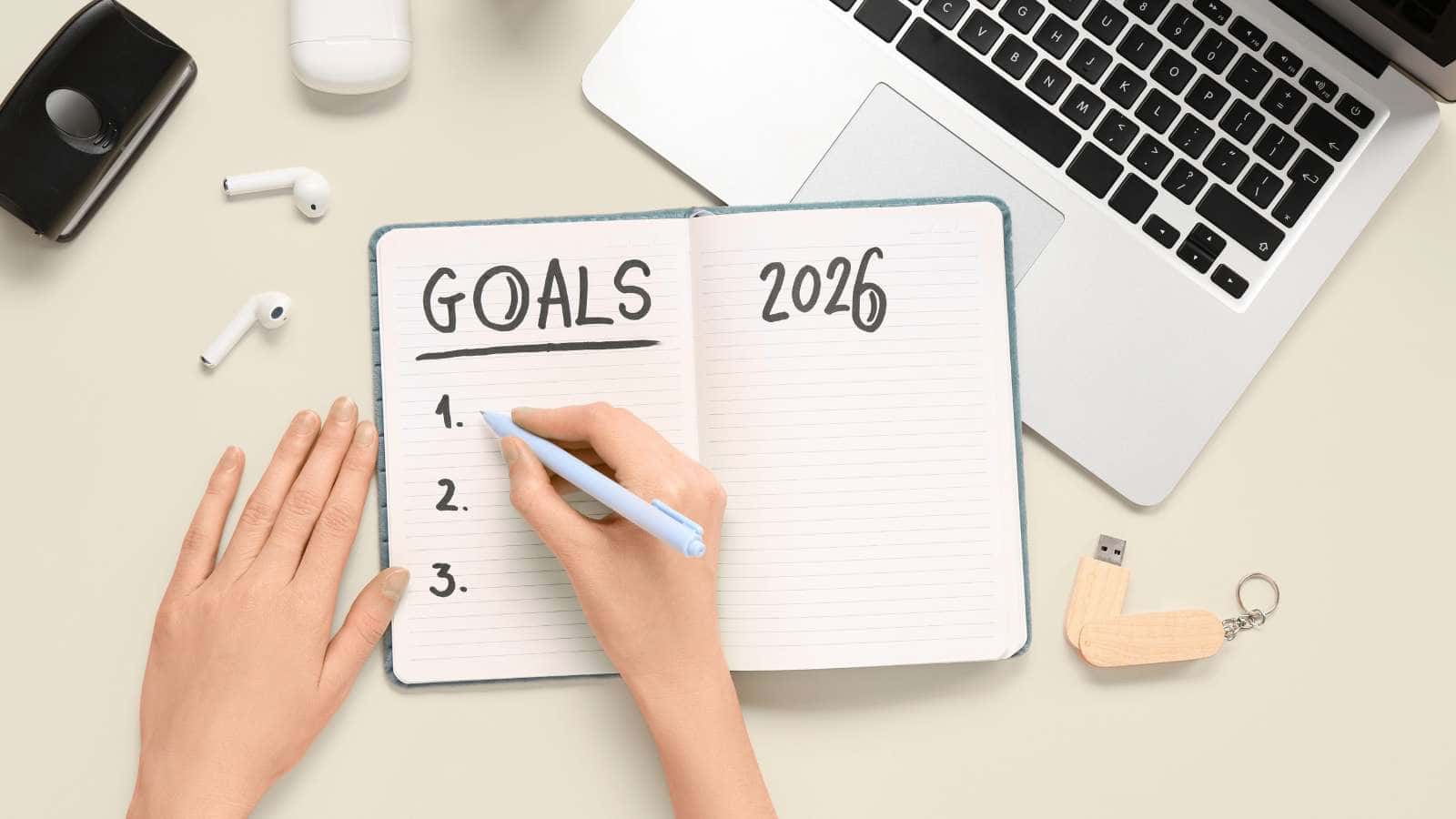 A person writes "GOALS 2026" in a notebook on a desk with a laptop, wireless earbuds, smartphone, pen, and USB drive.