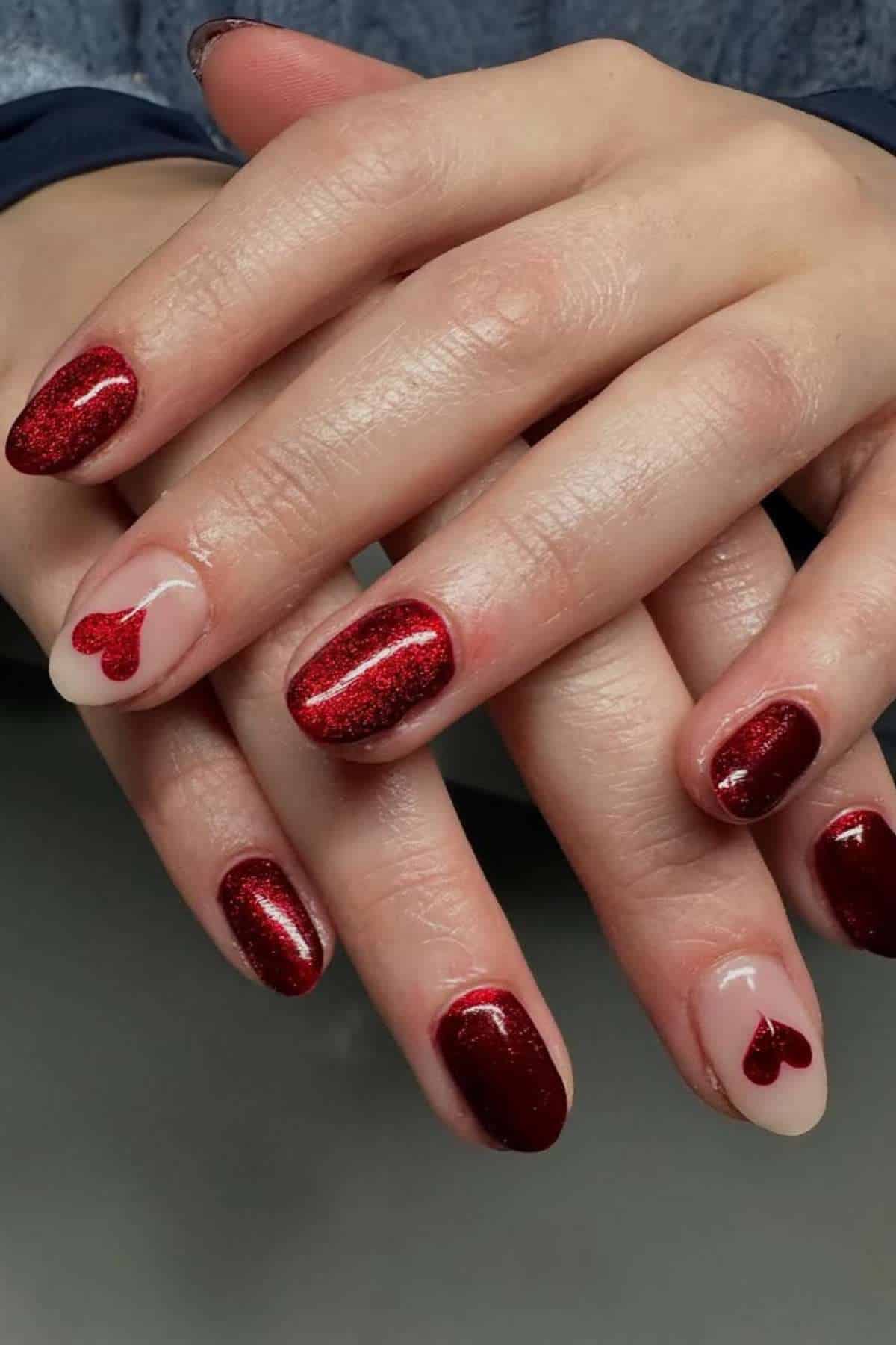 Hands with red glittery nail polish; ring fingers feature a nude base with small red heart designs near the cuticles.
