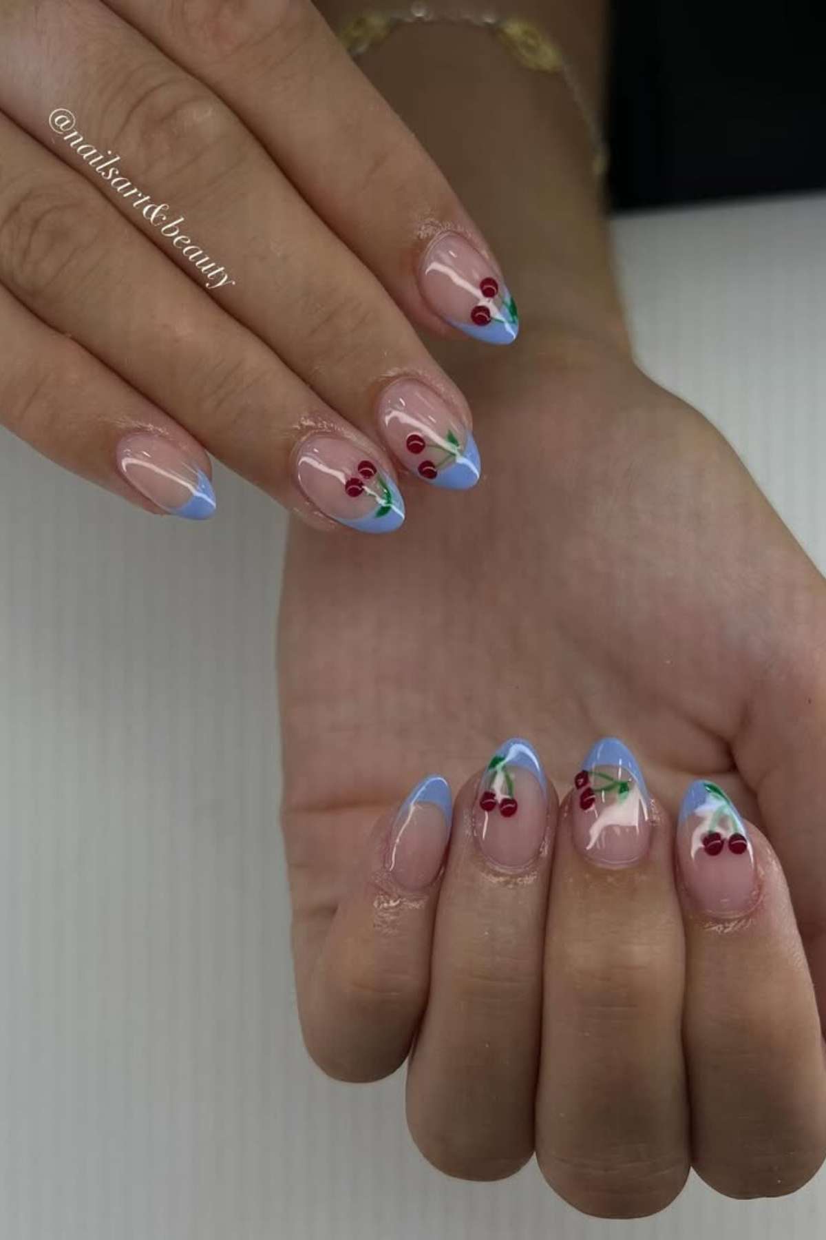 Hands with almond-shaped nails featuring light blue French tips and small red cherry designs on each nail.