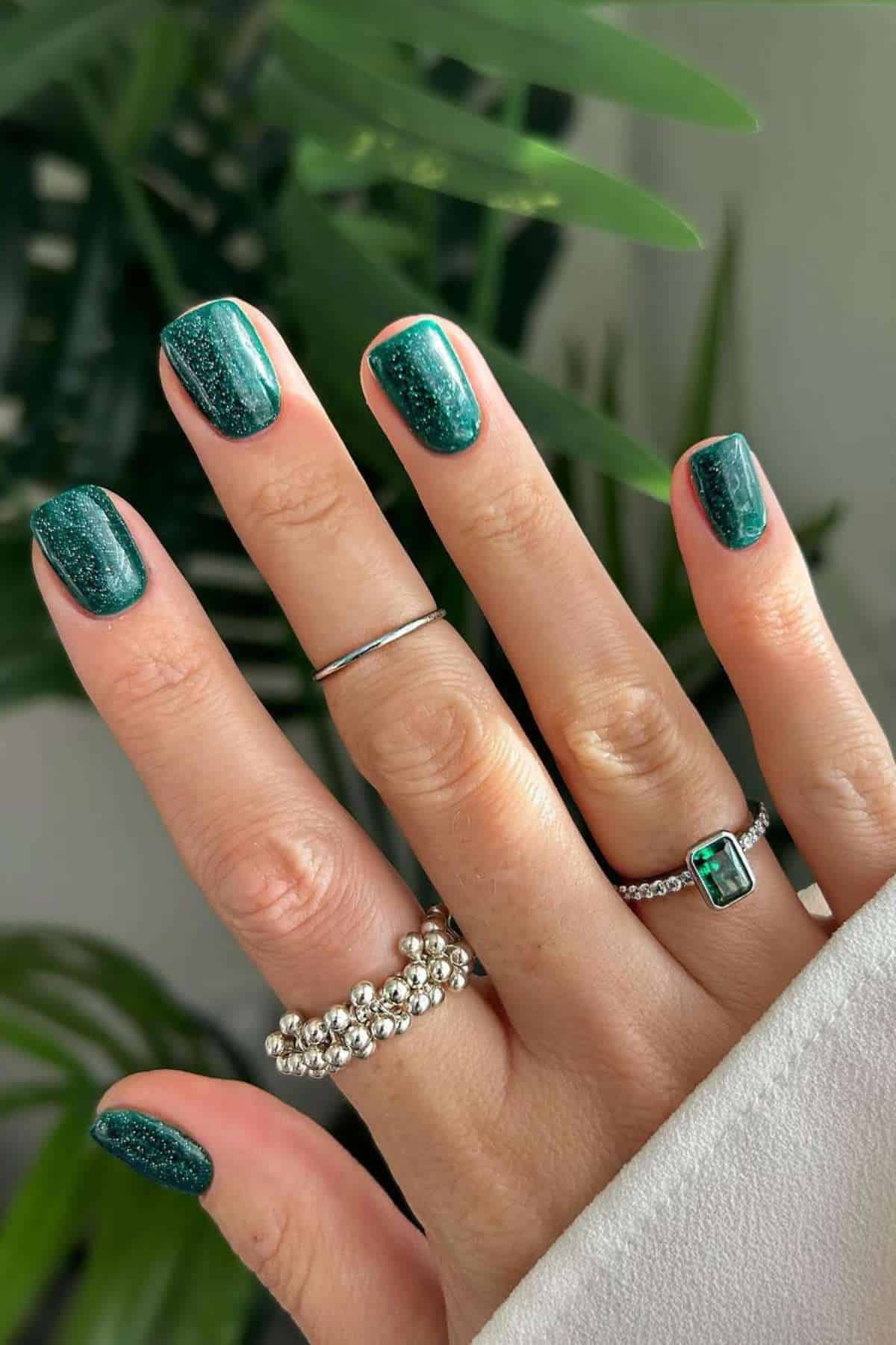 A hand with dark green glittery nail polish and three silver rings is shown against a leafy green background.