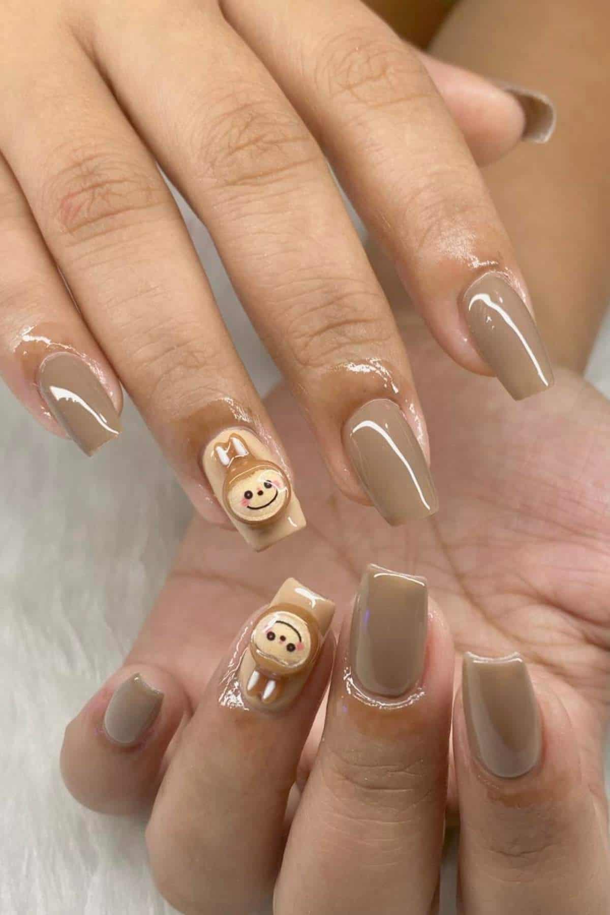 Hands with glossy, square-shaped nude nails; thumbs feature a small sloth face design on a beige background.