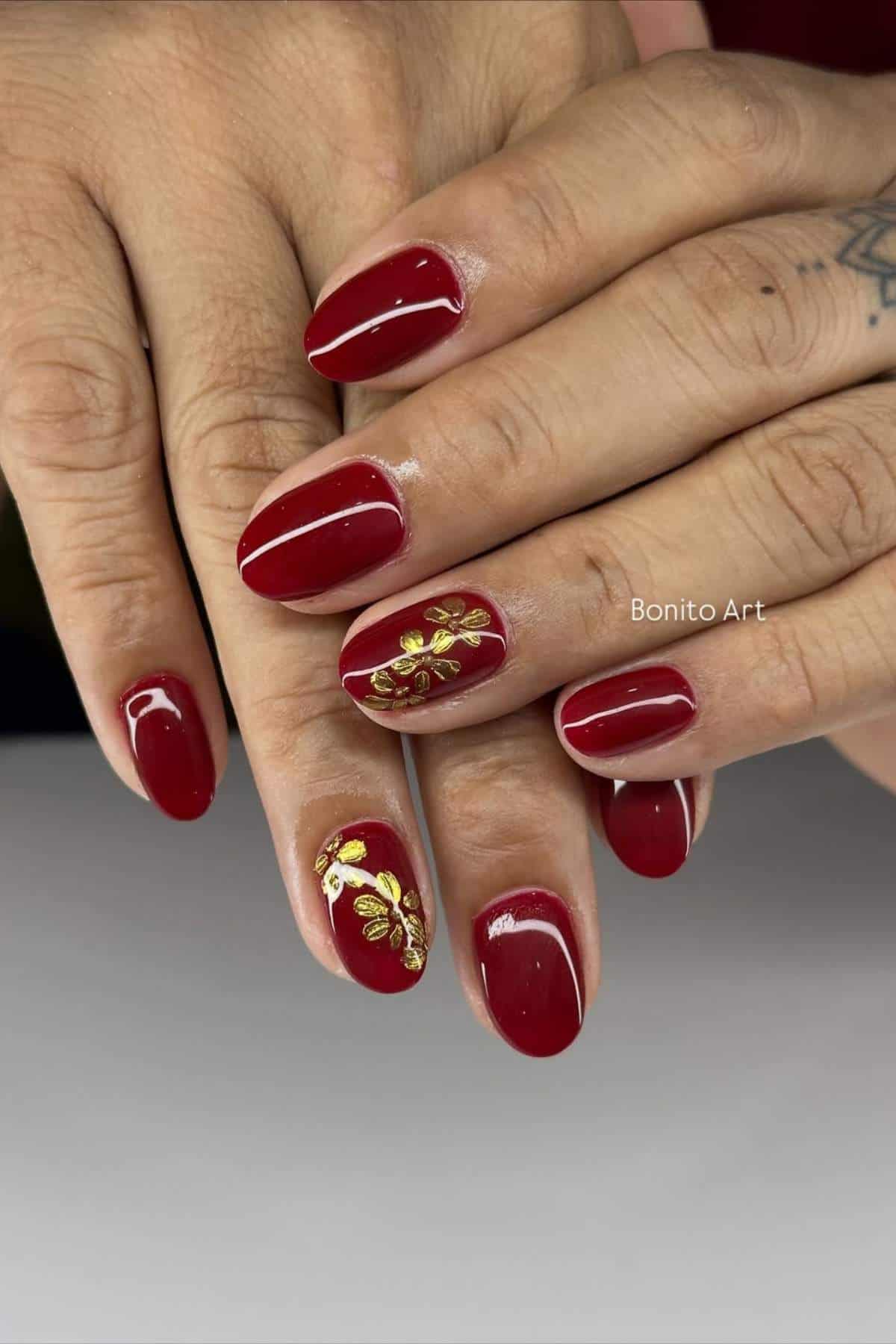 Hands with short, glossy red nails; gold floral designs are featured on the ring fingers.