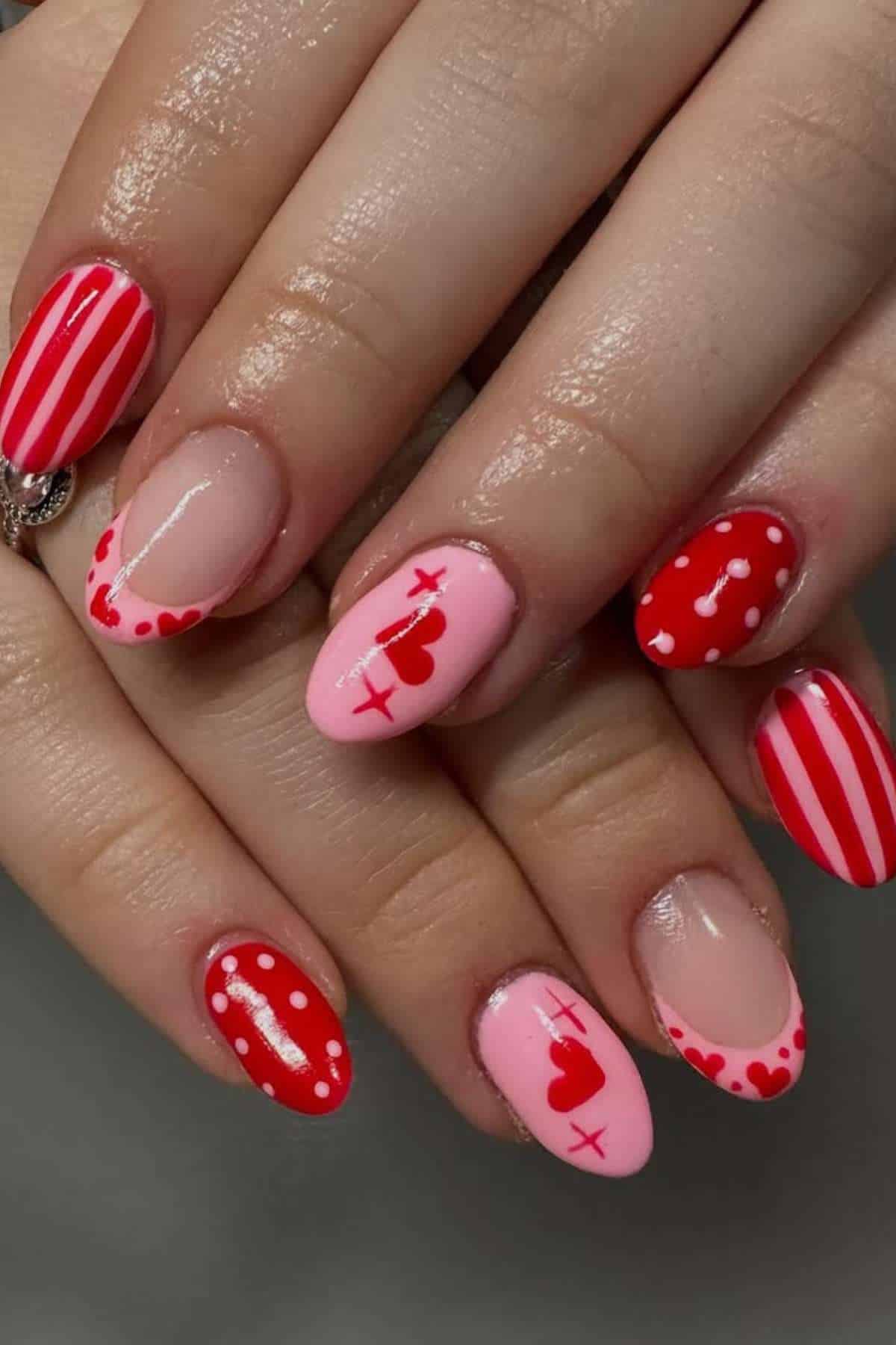 Hands with oval-shaped nails featuring red and pink designs, including stripes, dots, hearts, and X patterns on a neutral background.