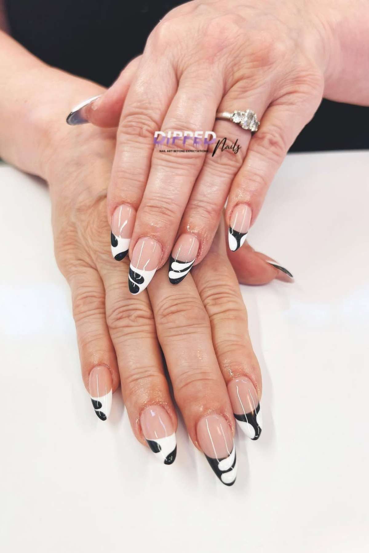 Hands with manicured nails featuring a black and white geometric French tip design. The person is wearing a silver ring with a clear stone on one finger.