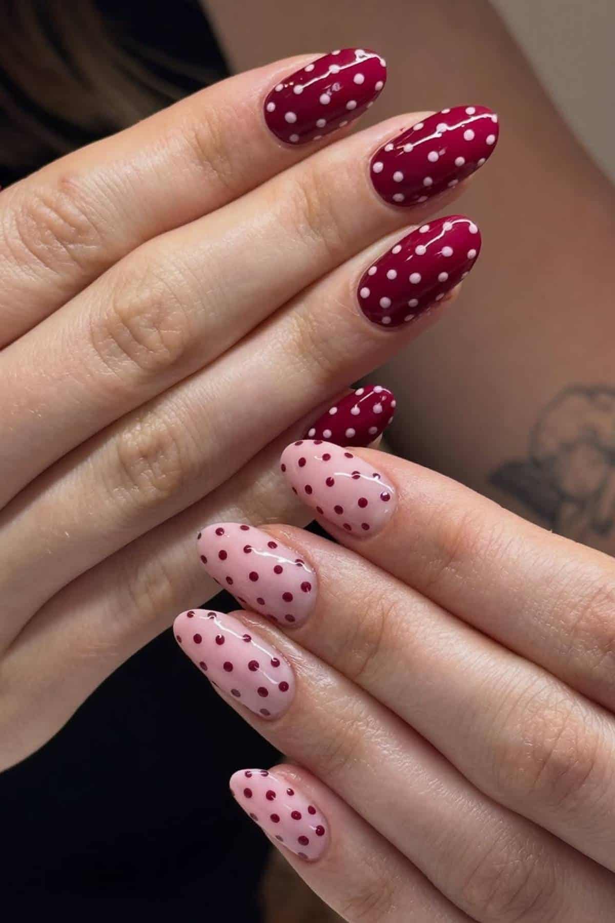 Two hands with manicured nails; one set painted maroon with white polka dots, the other light pink with maroon polka dots.