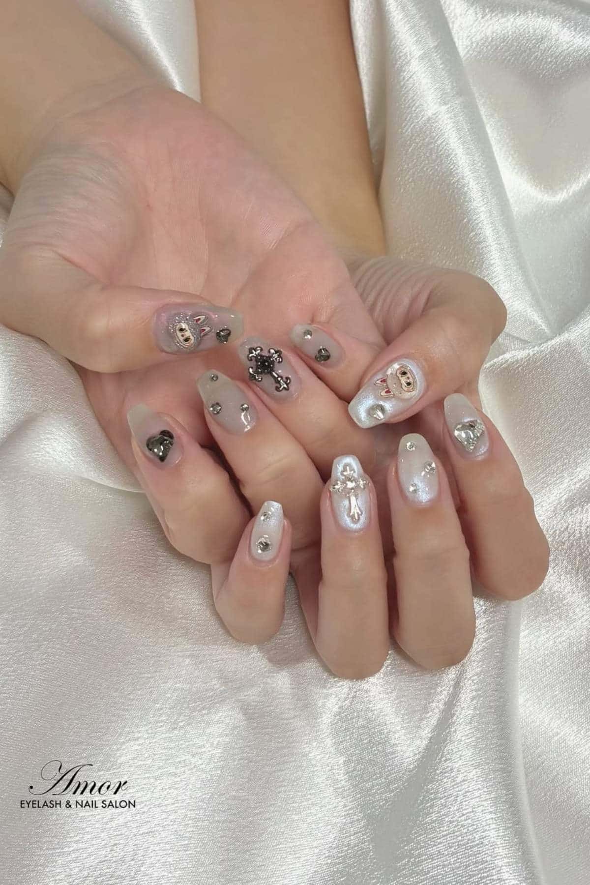 Hands with light-colored, glossy manicured nails featuring metallic and gem embellishments, various 3D designs, and small charms, shown against a white satin fabric background.