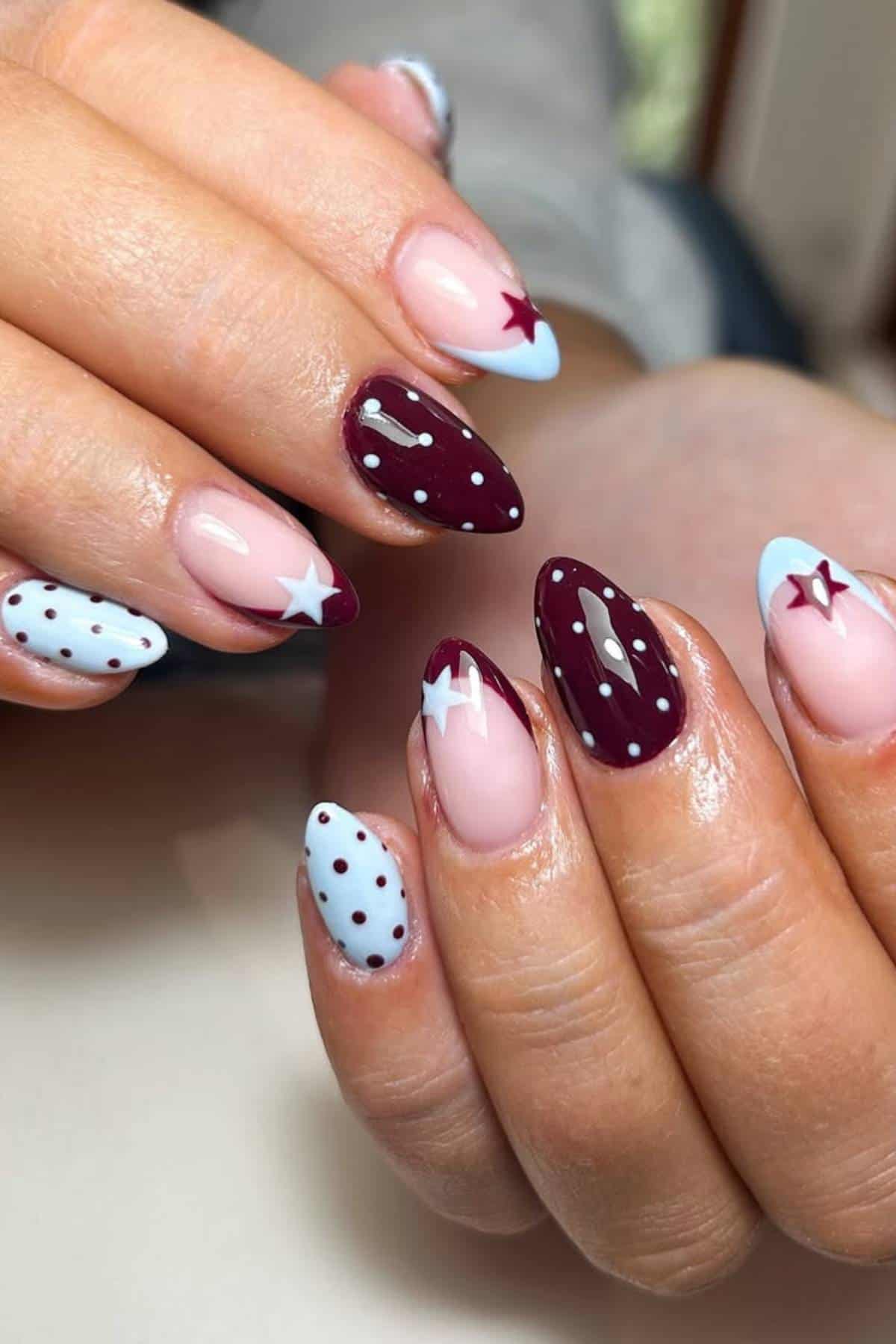 Hands with almond-shaped nails featuring a mix of burgundy, pale blue, and pink polish, decorated with stars and polka dot designs.