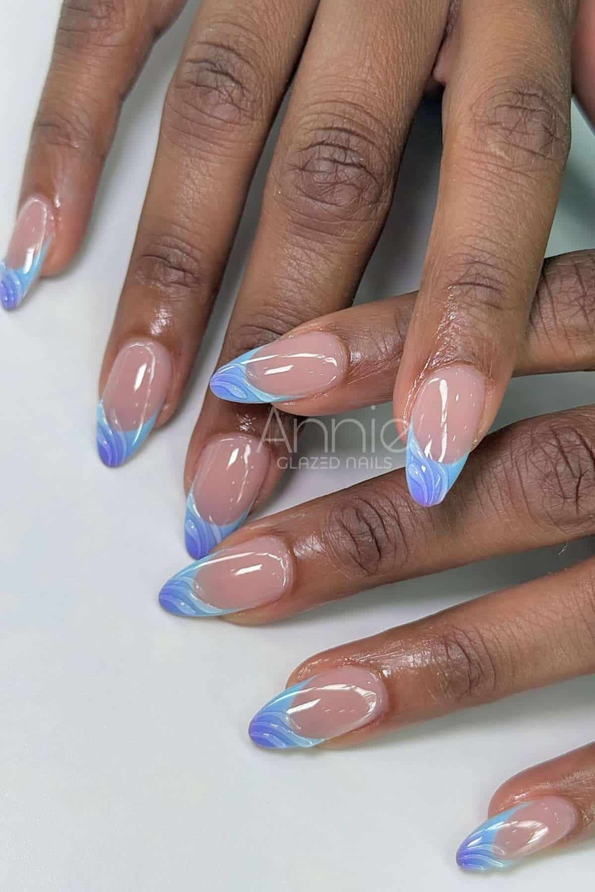 Hands with almond-shaped nails featuring a nude base and blue gradient French tips with a wavy design.