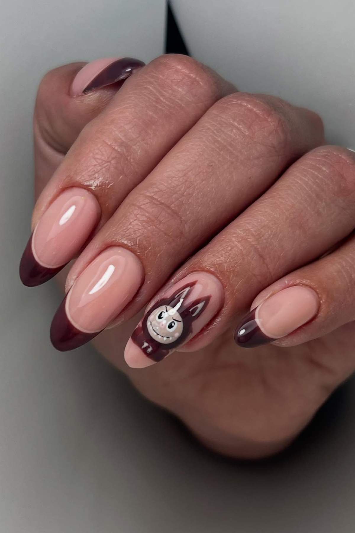 A hand with almond-shaped nails featuring a nude base, dark burgundy French tips, and a cartoon bunny design on the ring finger.