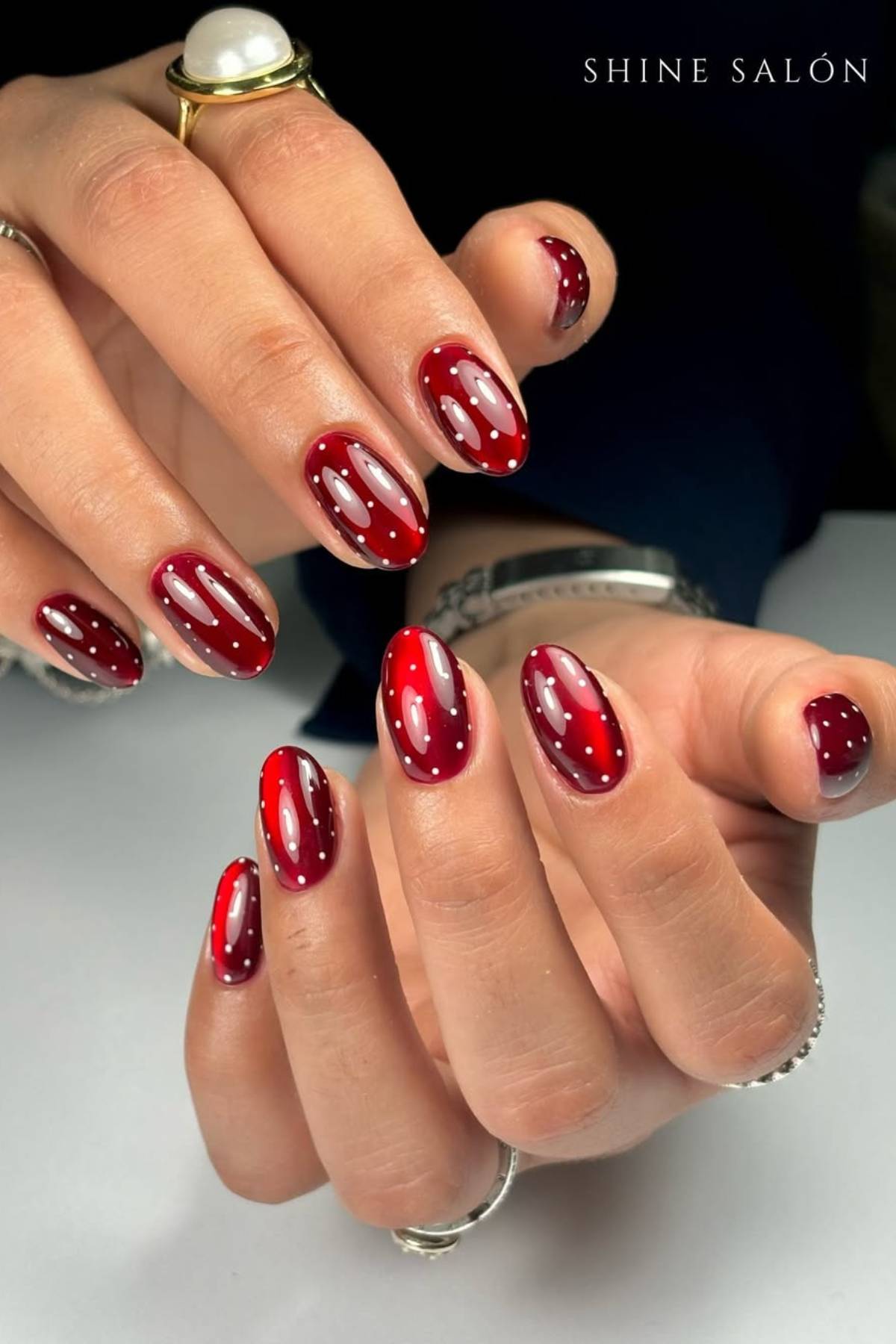 Close-up of hands with oval-shaped, glossy red nails featuring white dot and elongated highlight designs, adorned with rings.