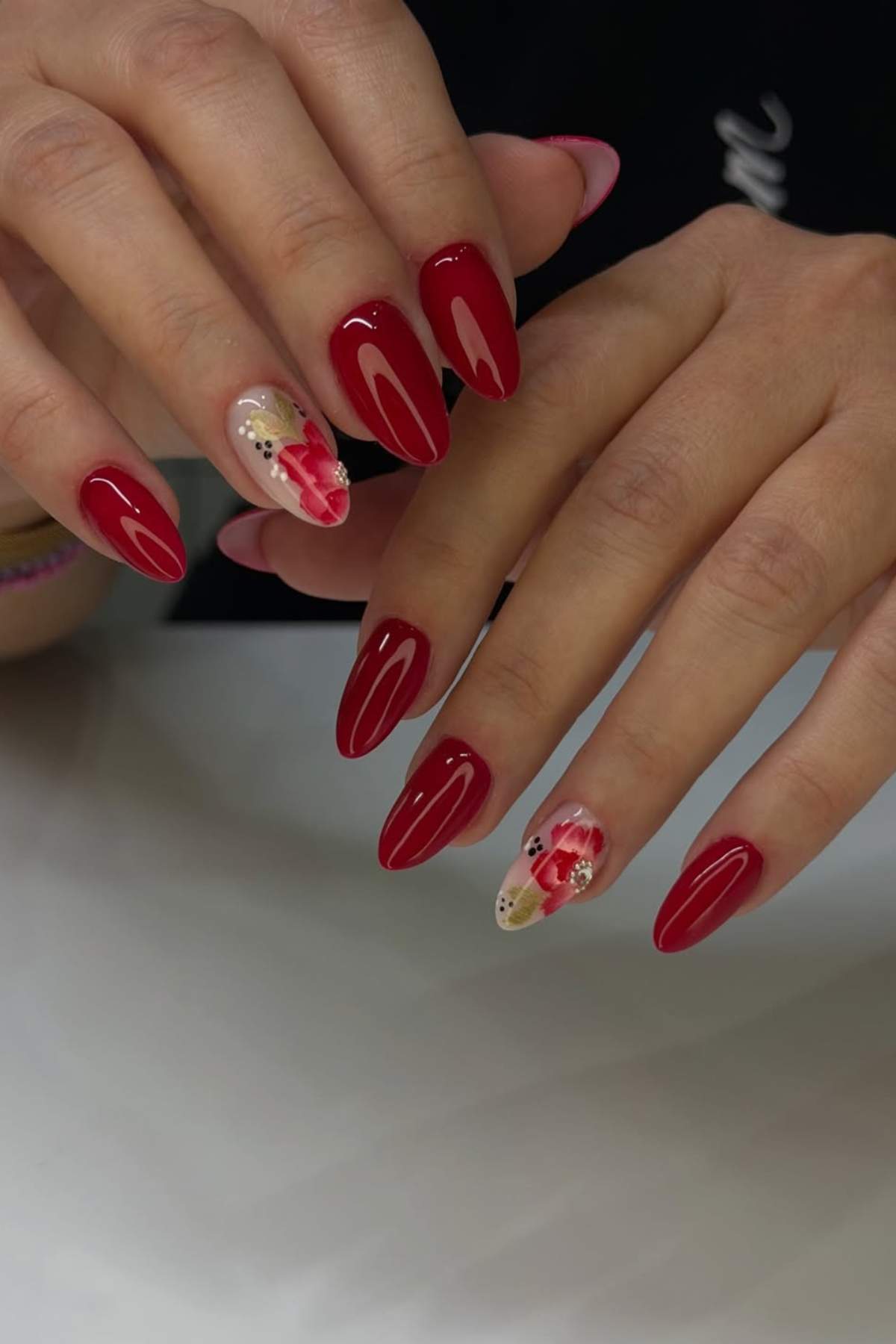 Hands with long, almond-shaped nails painted red; ring fingers feature floral nail art with pink and white flowers and gold accents.