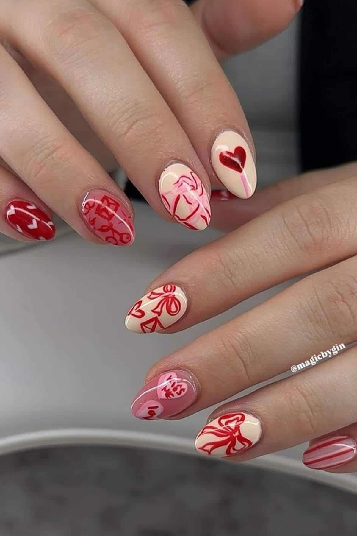 A hand with almond-shaped nails, painted in nude and pink tones, features red and pink abstract art, hearts, and floral designs on each nail.