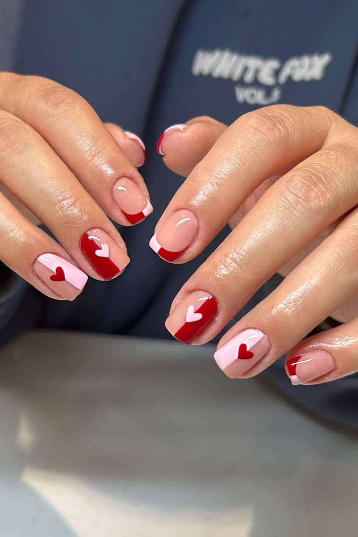 Close-up of hands with manicured nails featuring red and pink French tips, and white or red heart designs on a nude base.