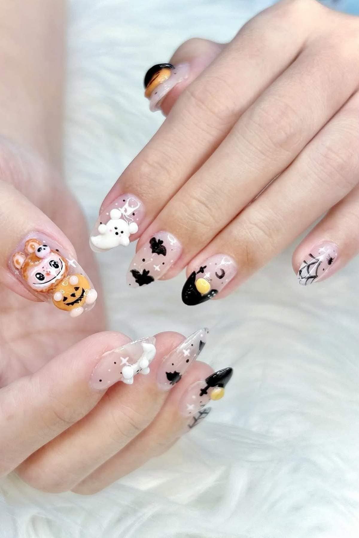 Close-up of hands with Halloween-themed nail art, featuring 3D decorations like pumpkins, ghosts, bats, spider webs, and bones on long, pointed acrylic nails.