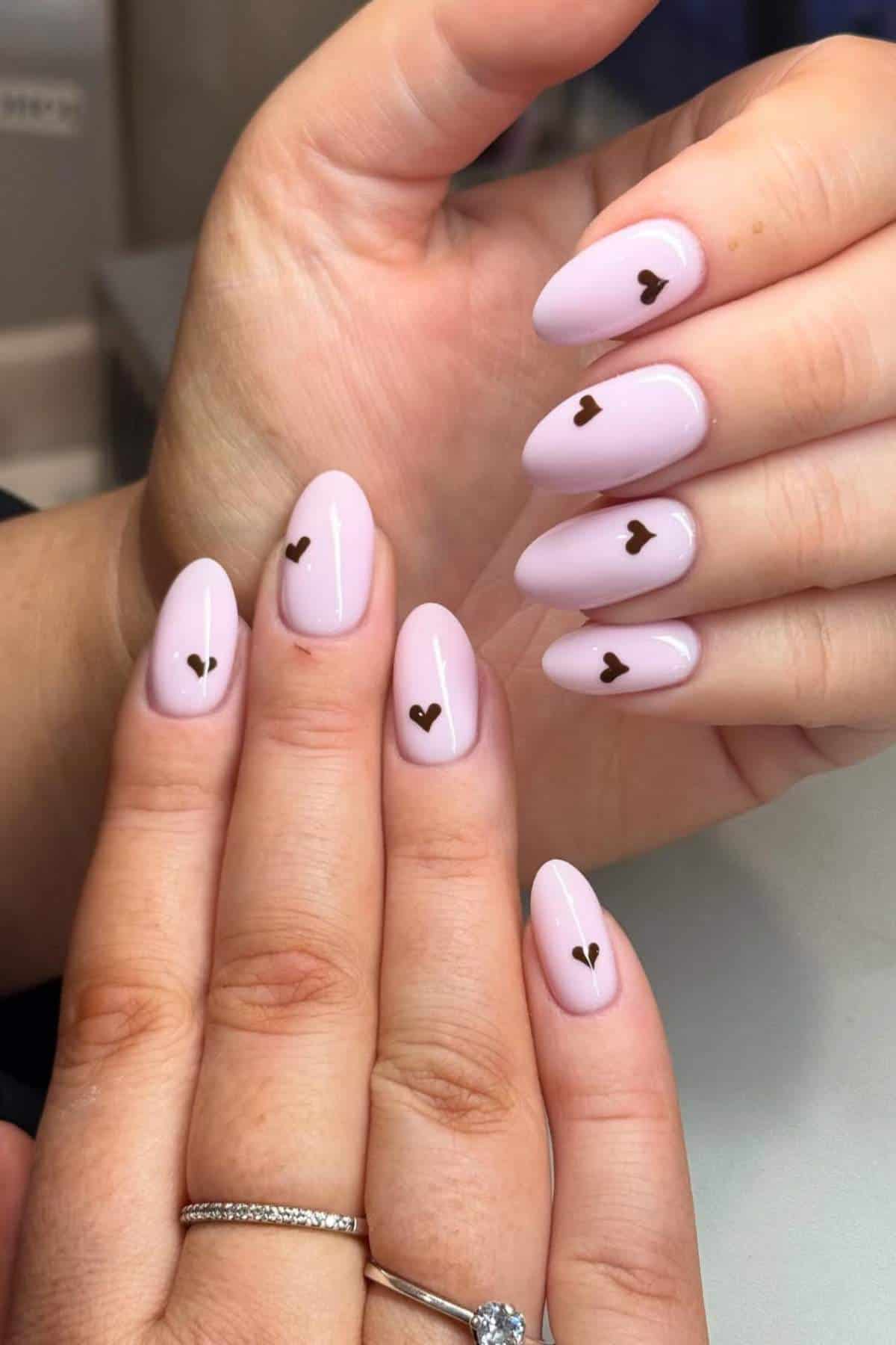 Close-up of hands with almond-shaped, light pink nails, each featuring a small black heart design near the cuticle.