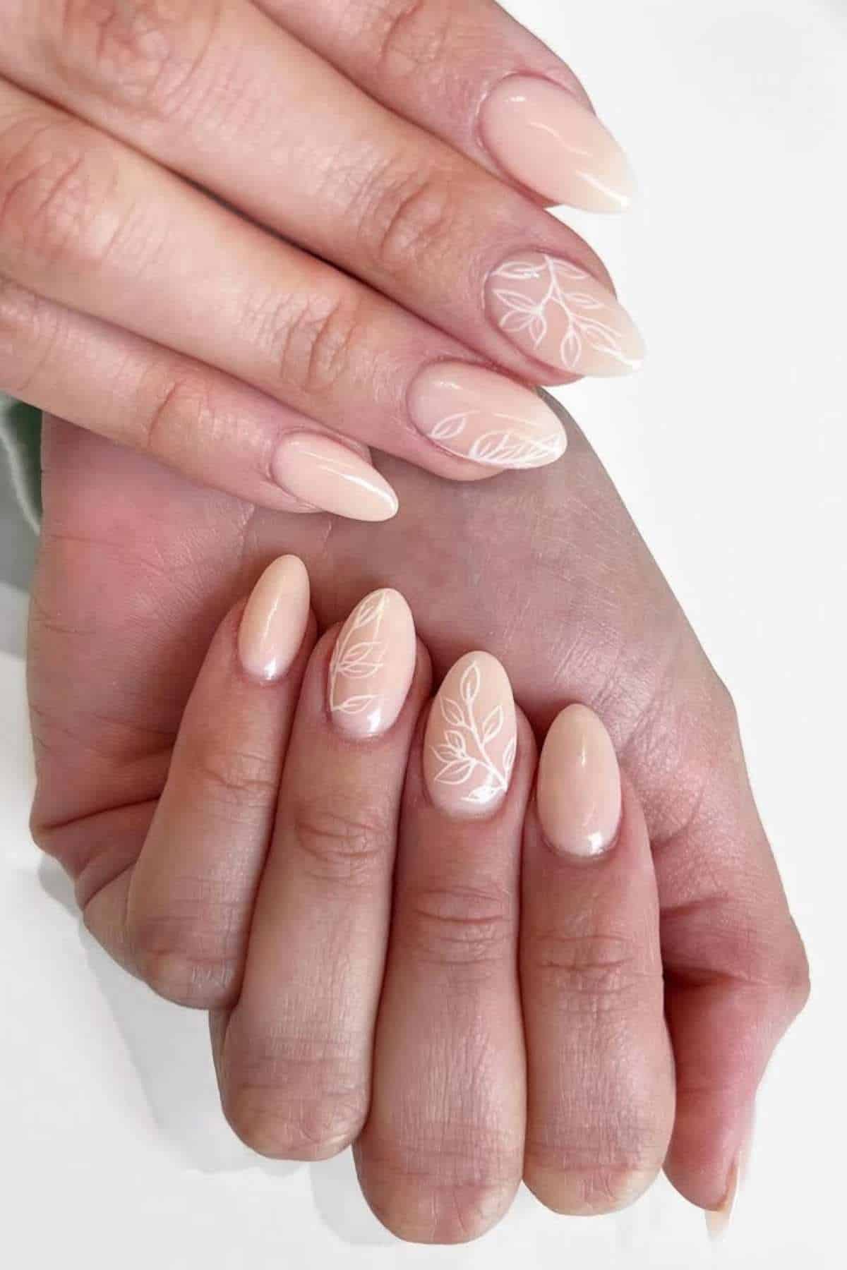 Hands with almond-shaped, nude-colored nails featuring minimalist white leaf designs on some fingers.