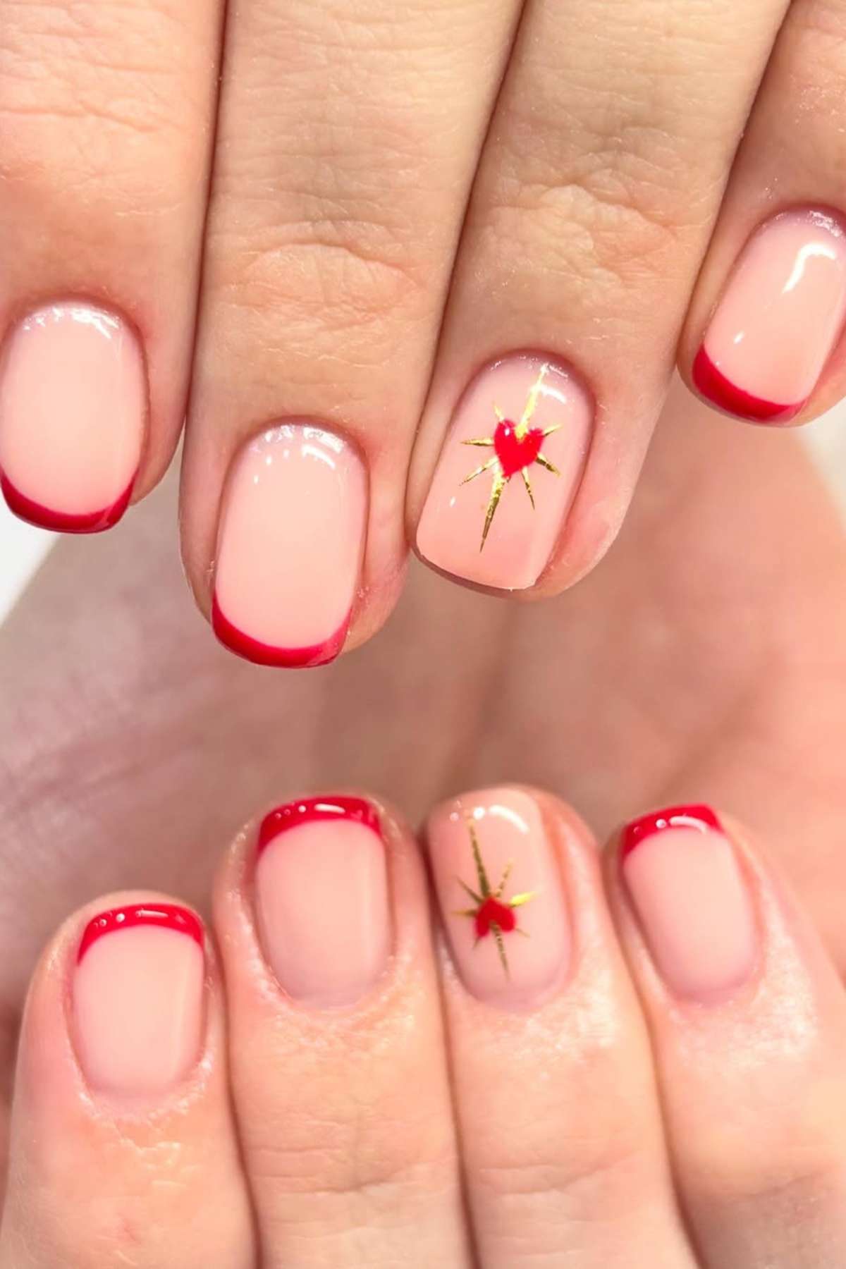 Hands with short, square-shaped nails painted with a pale pink base, red French tips, and a gold starburst and red heart design on the ring fingers.