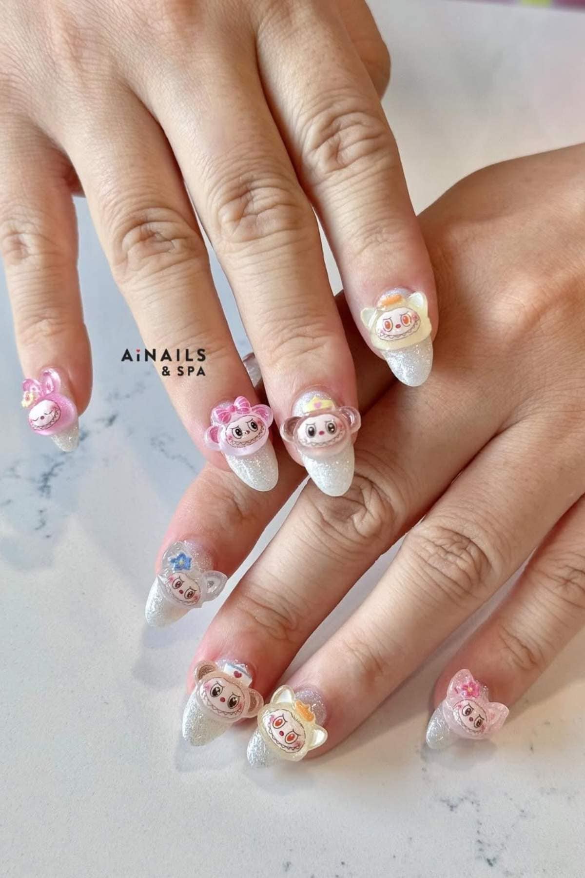 A set of manicured nails with white glitter polish and 3D animal face decorations in pink, cream, and brown tones on each nail.