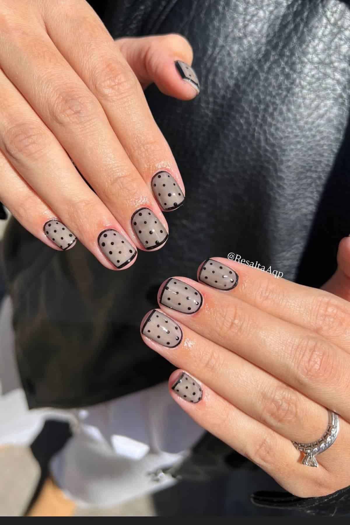 Close-up of hands with nails painted in a sheer nude color, black polka dots, and black French tips. The person wears a silver ring and a black leather jacket.