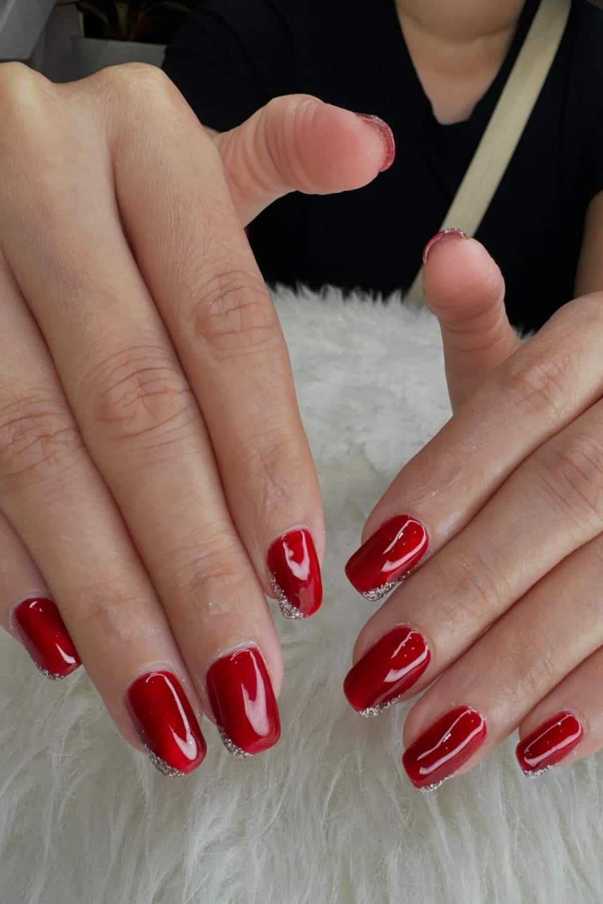 Close-up of hands with freshly painted red nails featuring a silver glitter accent along the cuticles, resting on a white furry surface.