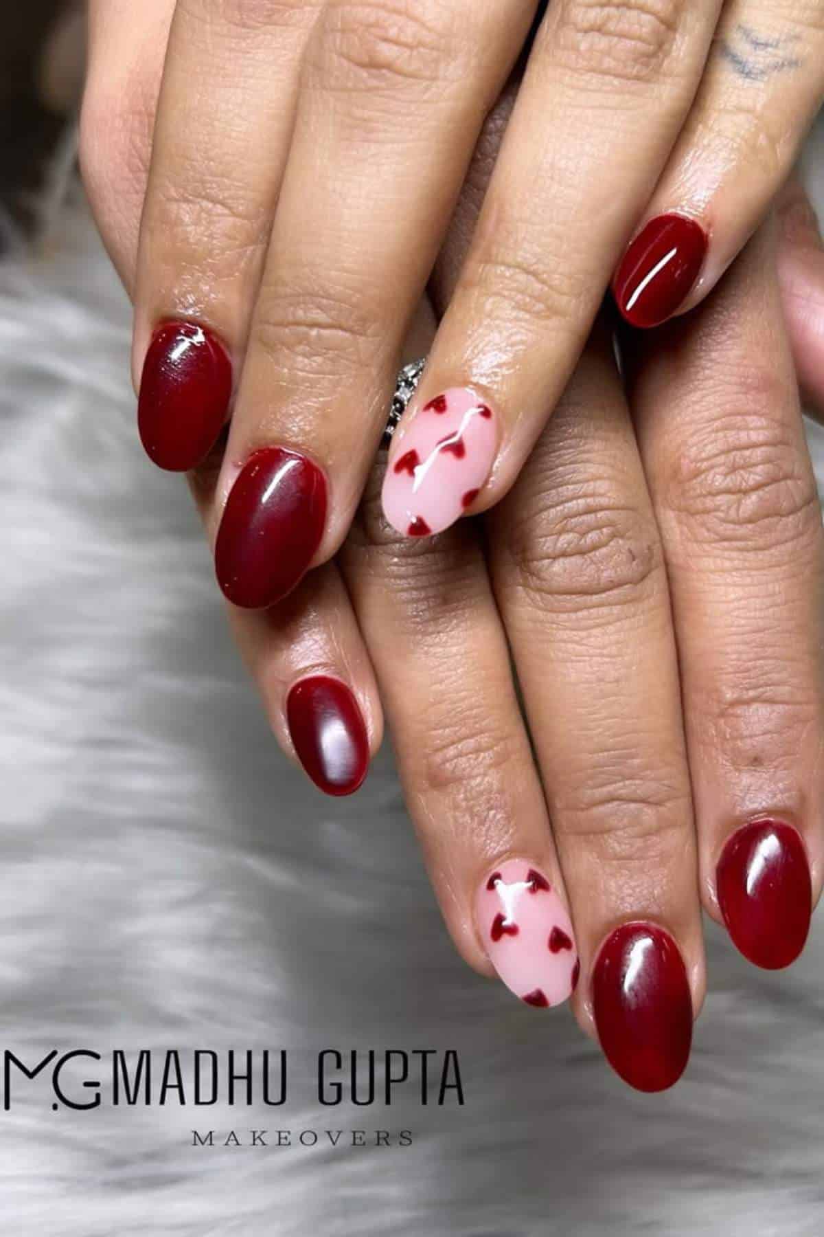 Hands with red oval-shaped nails and two accent nails painted light pink with small red heart designs. Logo "MG Madhu Gupta Makeovers" is visible at the bottom.