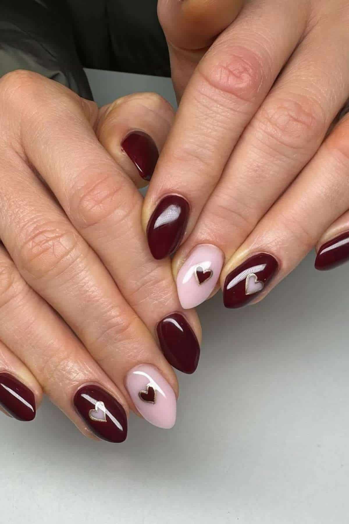 Hands with almond-shaped nails painted burgundy and pale pink, featuring small metallic heart-shaped nail art on some nails.