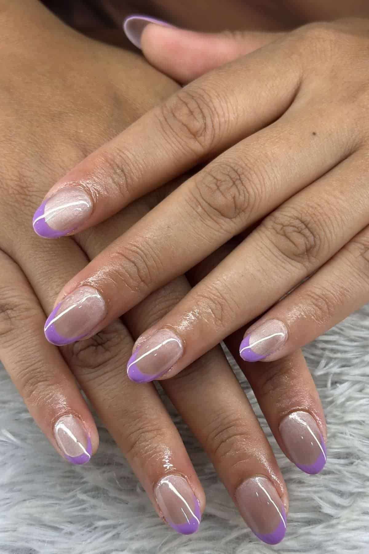 Close-up of hands with natural nails featuring a glossy nude base and light purple French tips, displayed on a soft, gray textured surface.