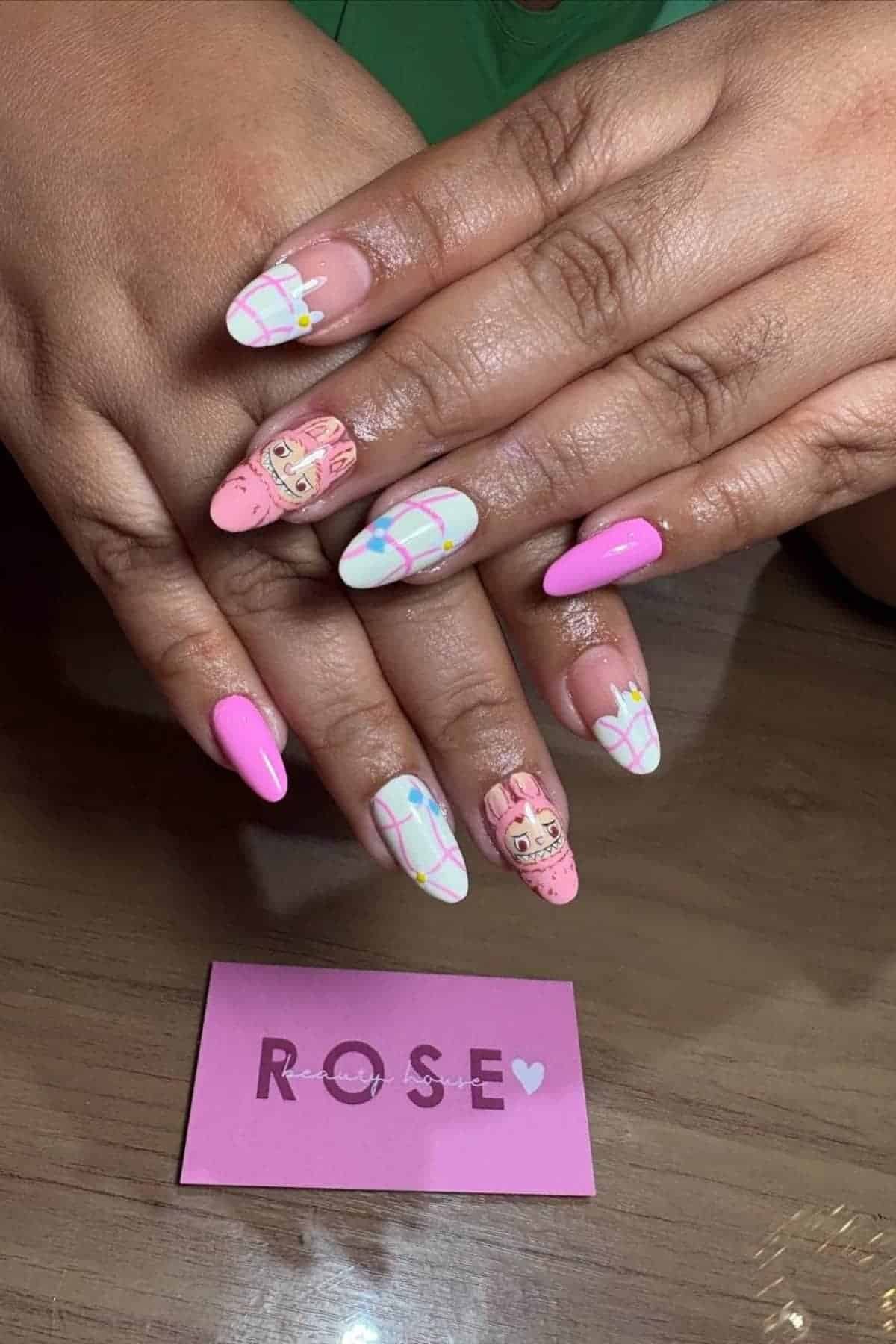 Hands with long, almond-shaped nails featuring pink, white, and pastel plaid designs, with cartoon character art on two nails. Pink card labeled "ROSE" lies on the table below.
