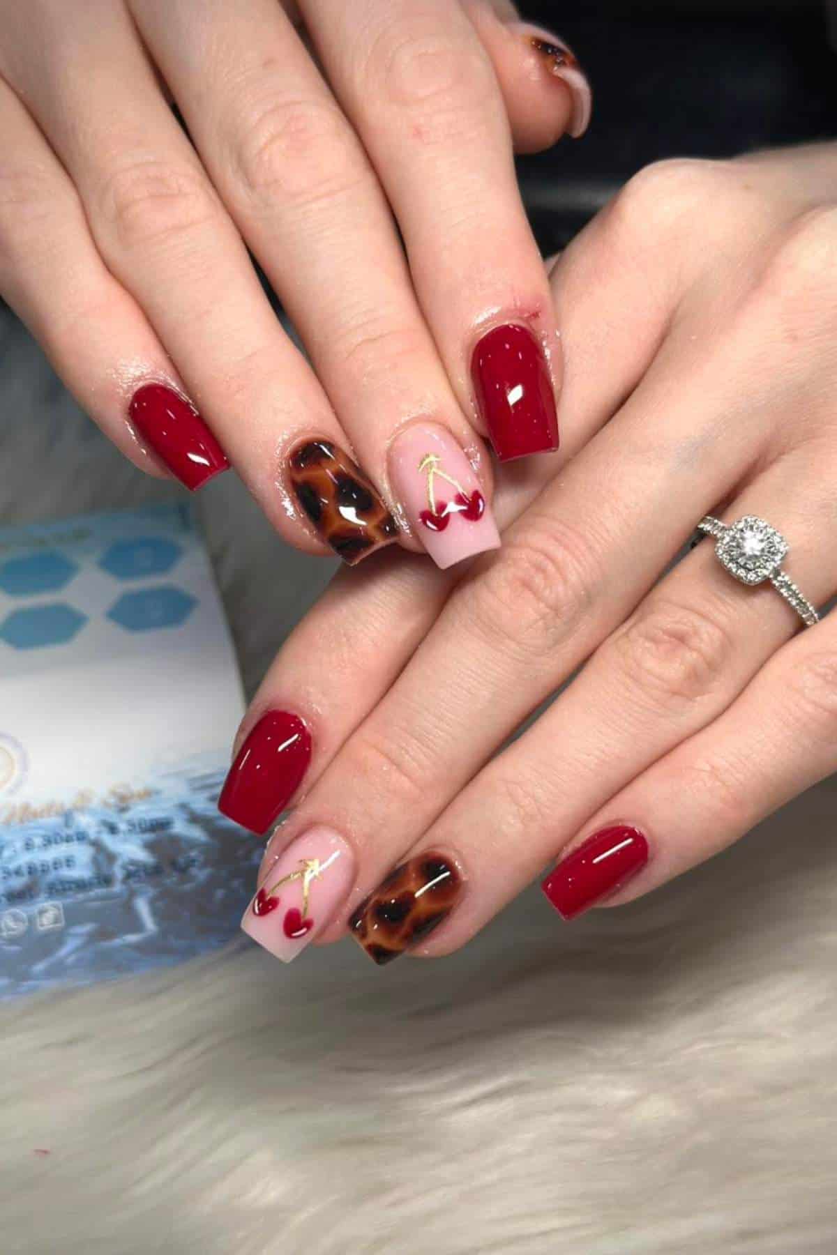 Hands with manicured nails featuring red polish, tortoiseshell pattern, and accent nails with cherry designs; a diamond ring is worn on one finger.