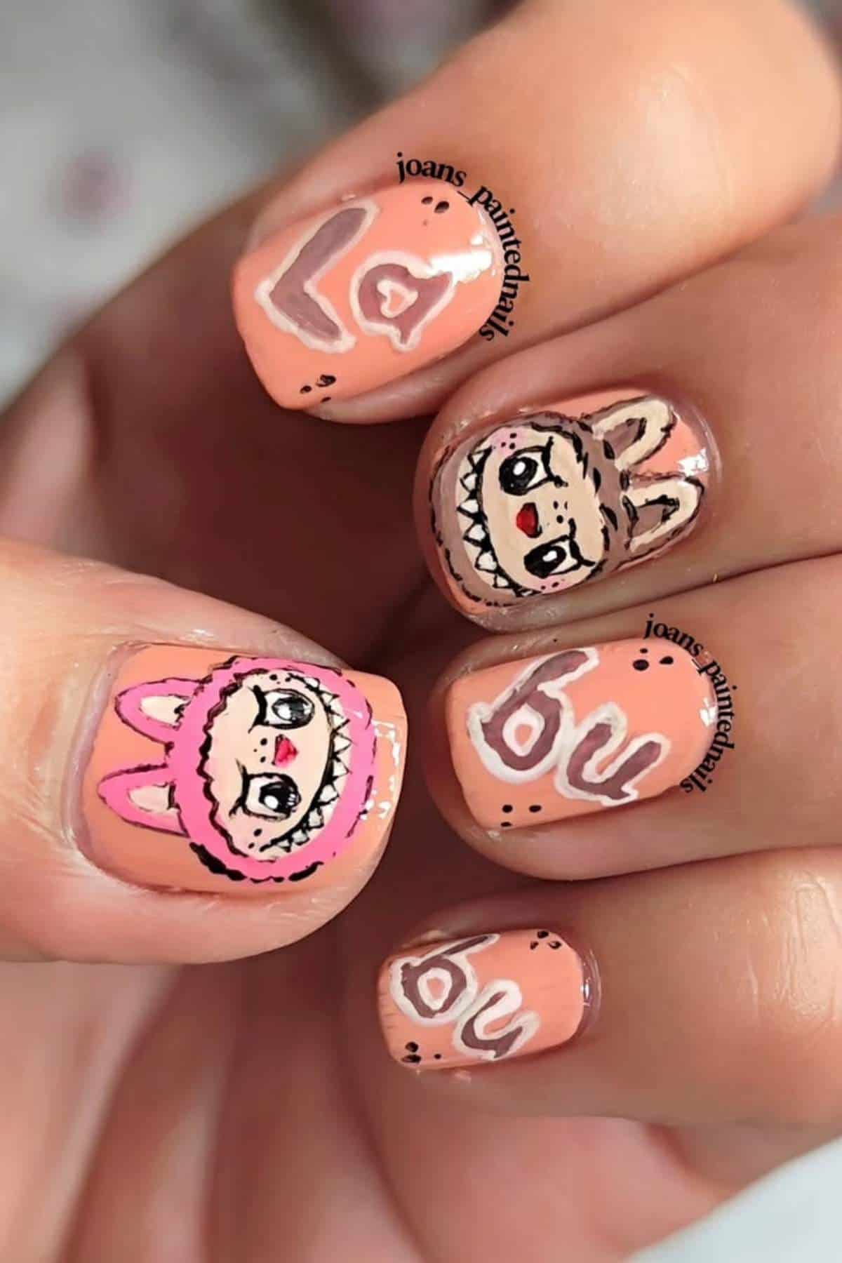 Close-up of fingernails painted with peach-colored polish, featuring cartoon faces in bunny hoods and the words "la bu" in white lettering on the nails.