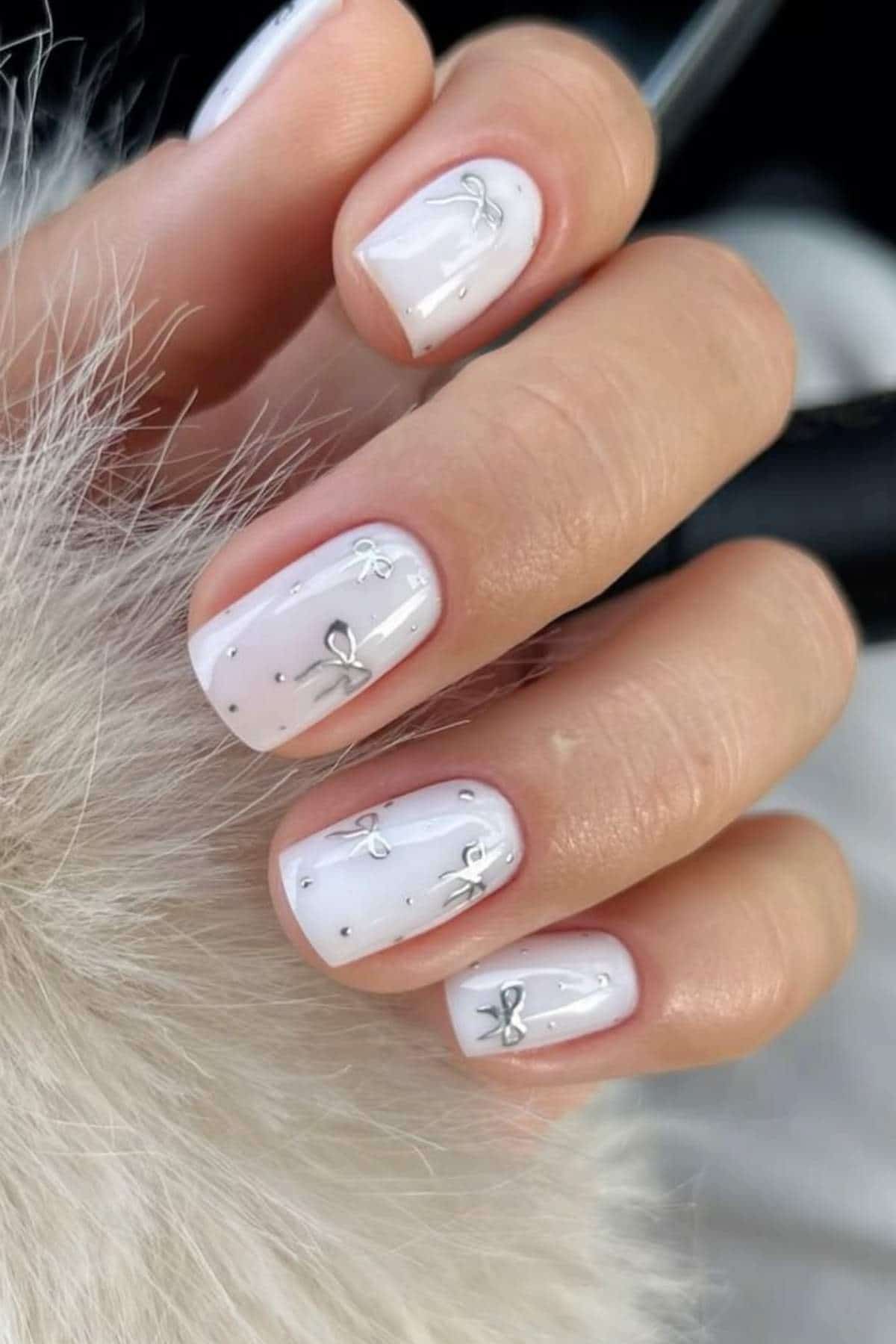 A hand with short, square-shaped nails painted pale pink with small silver bow designs and tiny black dots on each nail.