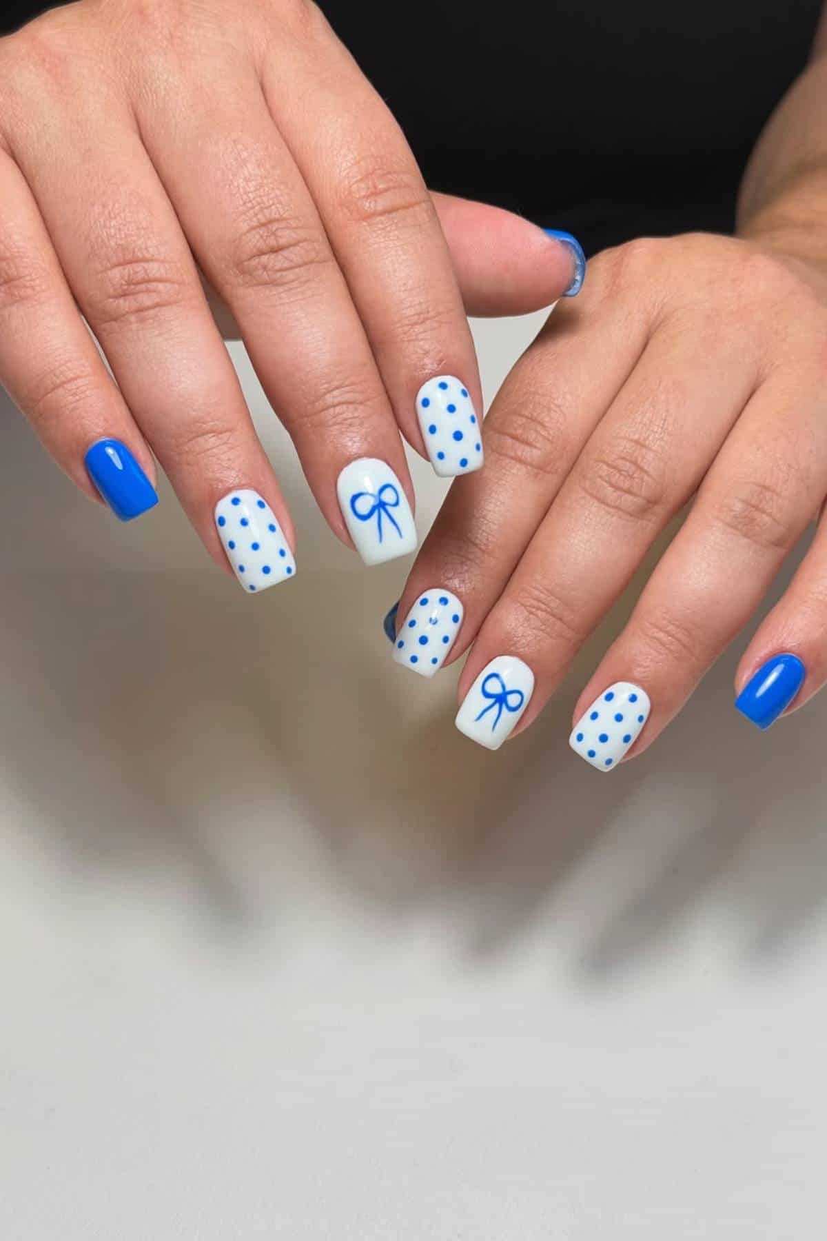 Hands with blue and white nail art; white nails feature blue polka dots and bows, while other nails are painted solid blue.