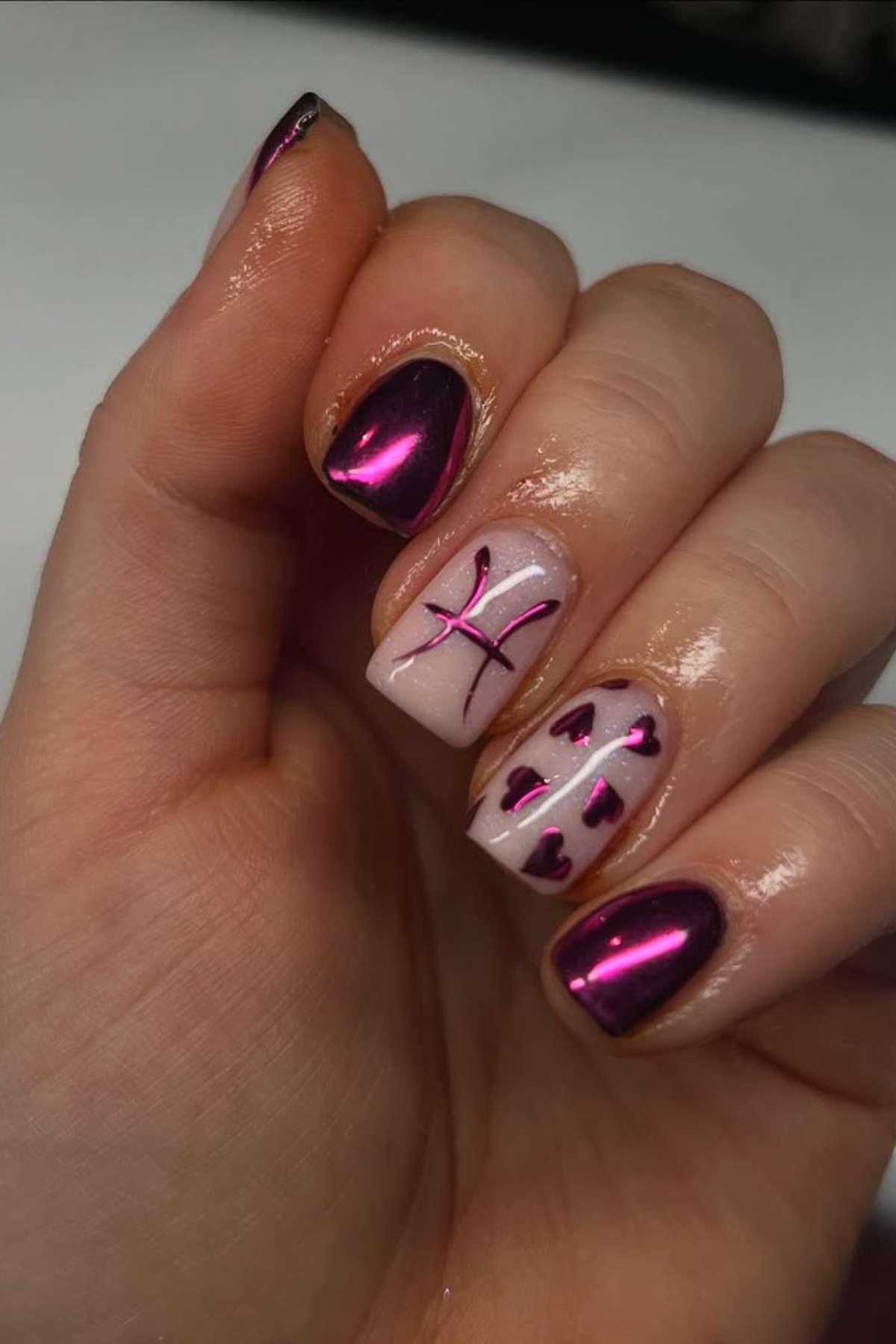 A hand with metallic magenta nail polish; the ring finger has a Pisces symbol and the middle finger has small magenta hearts on a light base.