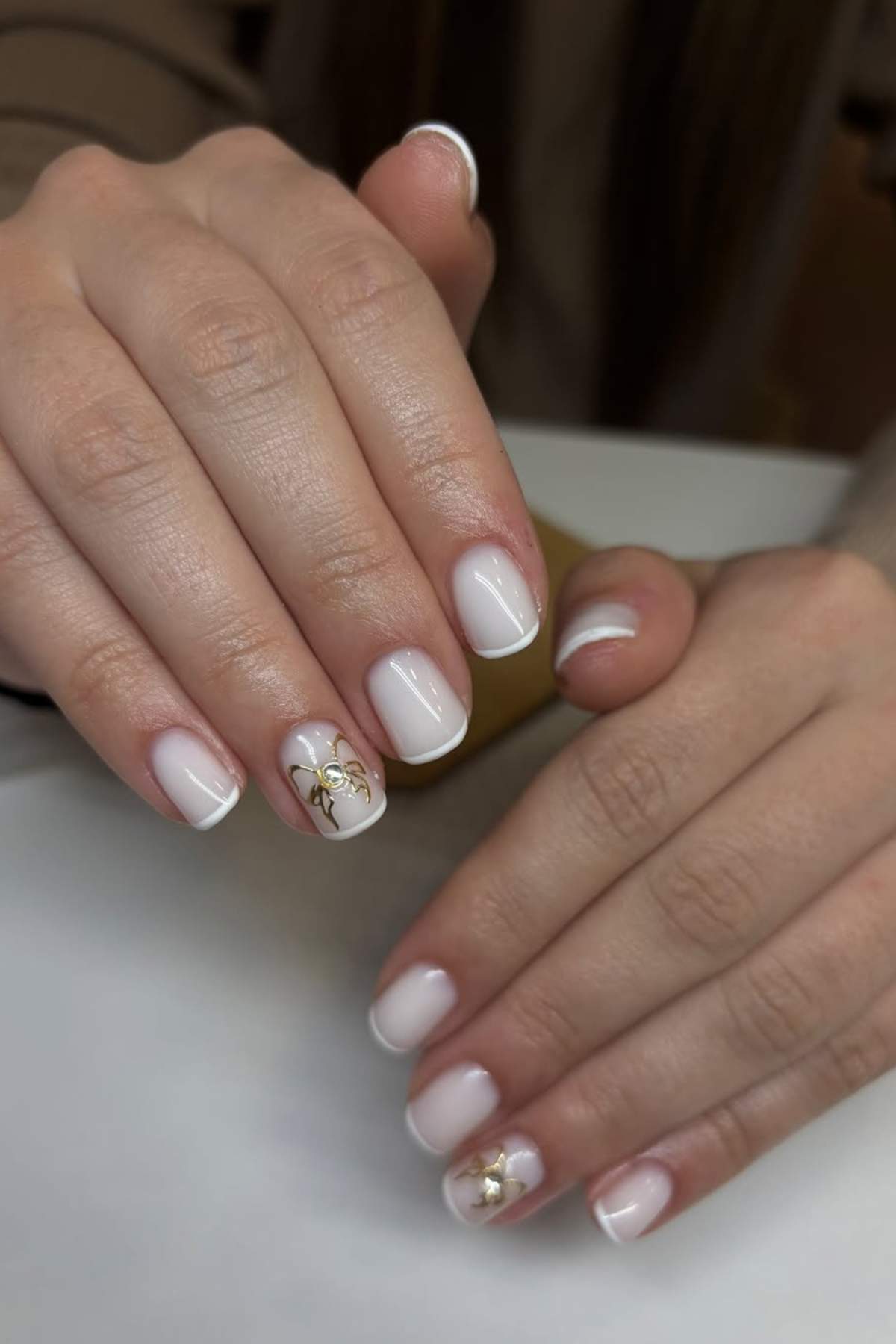 Close-up of hands with short, square-shaped nails painted in pale pink and white, featuring gold floral designs on two accent nails.