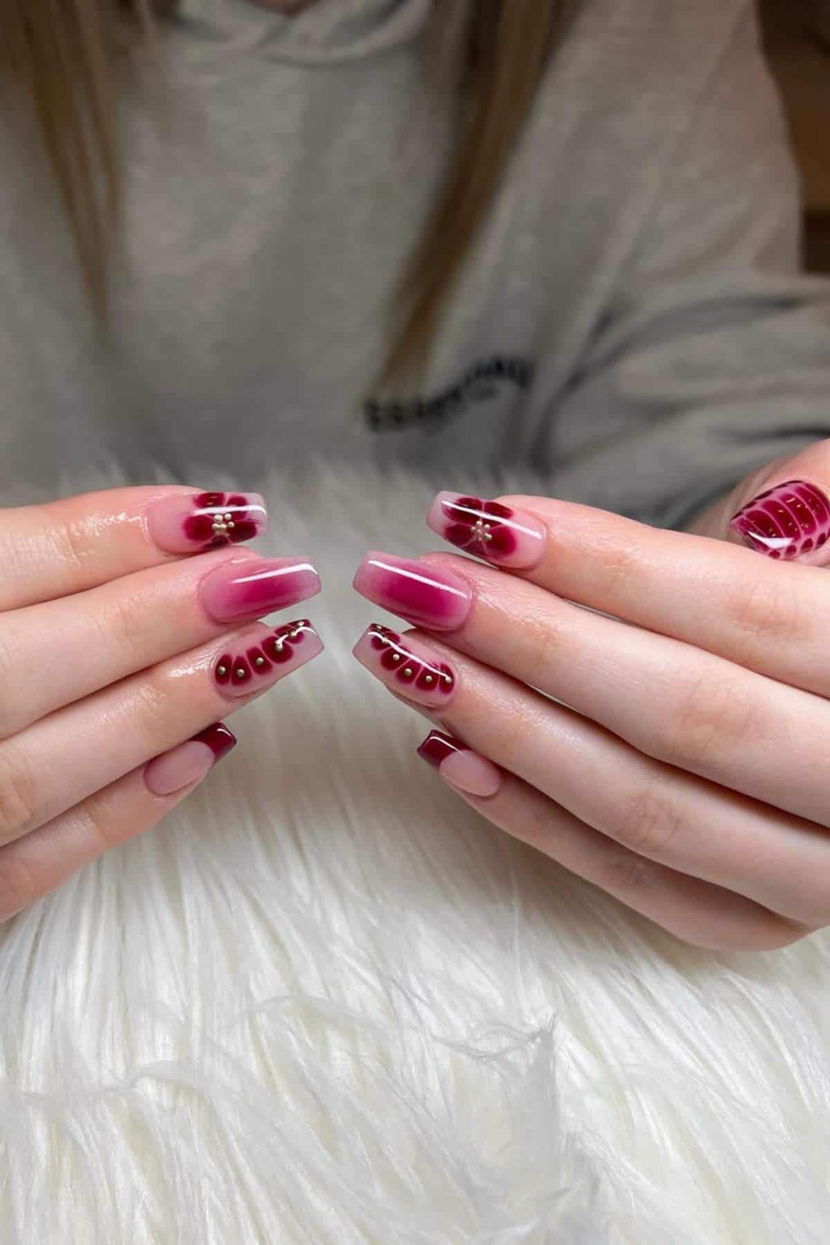 Hands with long square acrylic nails featuring red and pink ombre, flower designs, and dotted patterns, displayed on a white furry surface.