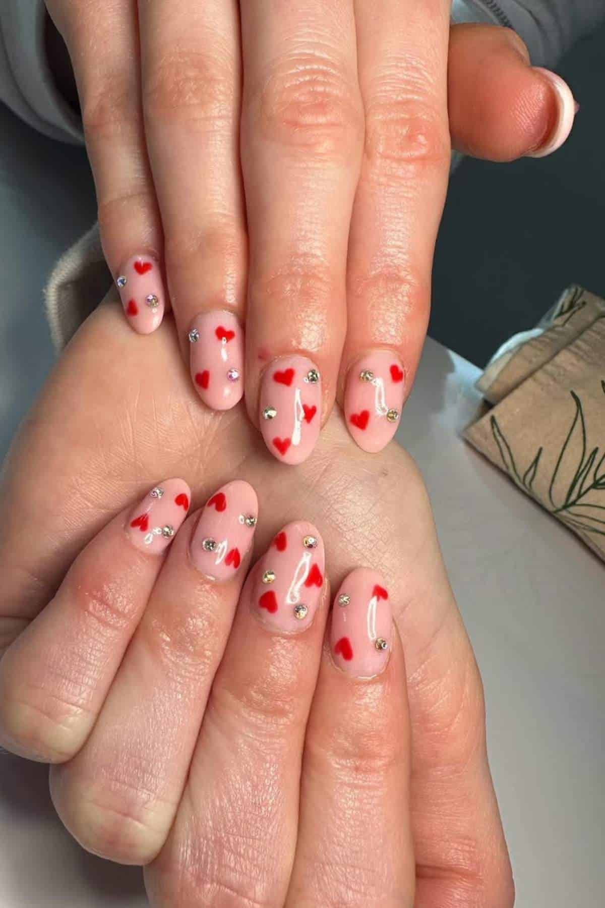 Hands with almond-shaped nails painted in a nude color, decorated with small red hearts and silver rhinestones.