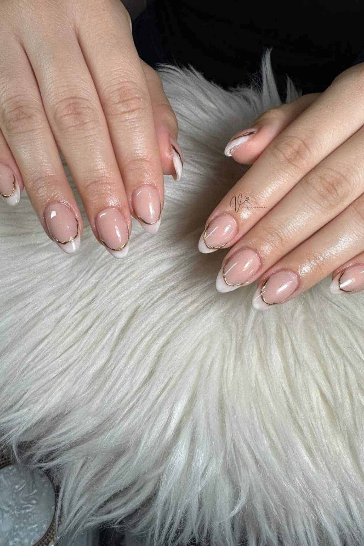 Two hands with almond-shaped nails featuring a nude base and white French tips outlined with a thin gold line, resting on a white, fluffy surface.