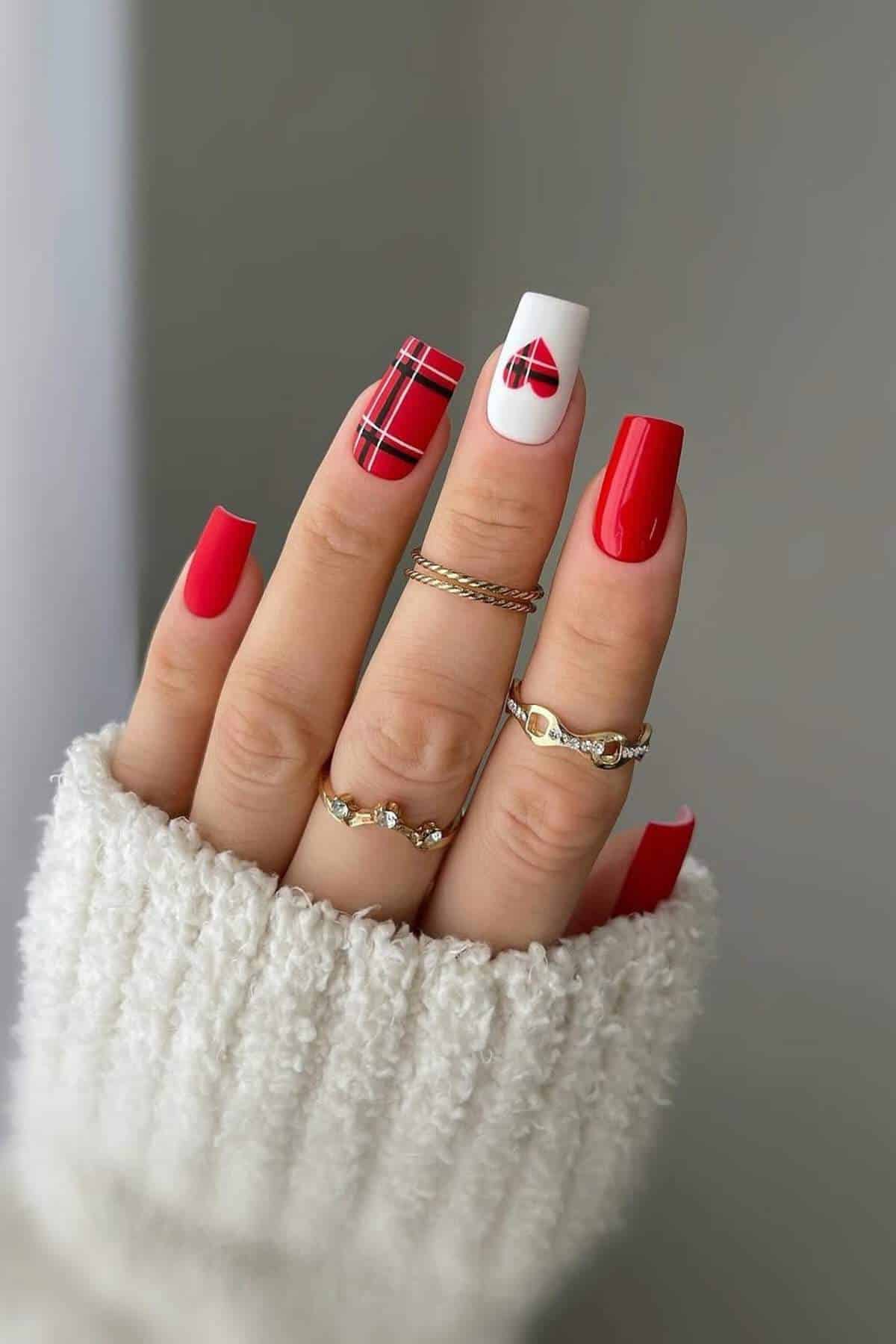 A hand with red and white square-shaped nails, featuring plaid and bow designs on two accent nails, wearing gold rings and a white knit sweater.