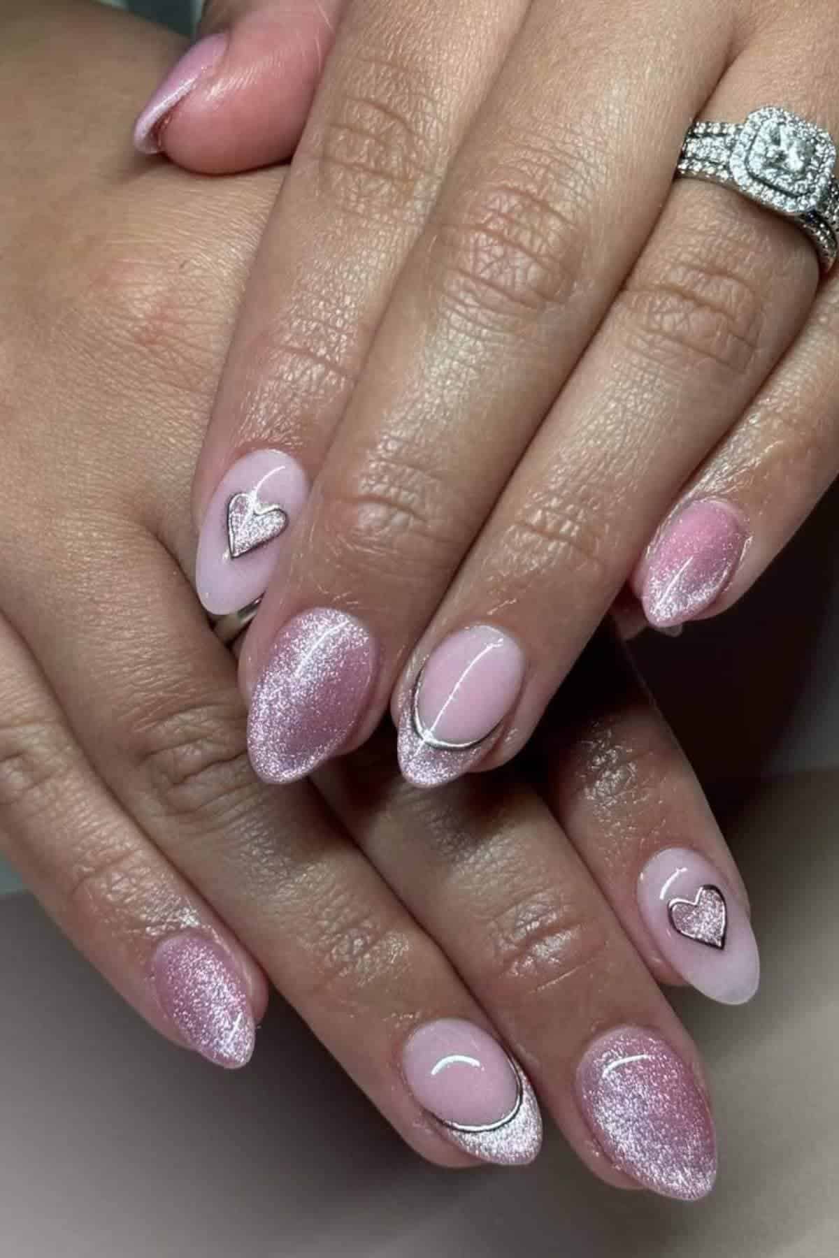 A close-up of hands with almond-shaped pink gradient nails, accented with metallic designs and heart-shaped embellishments. The person is wearing a diamond ring.